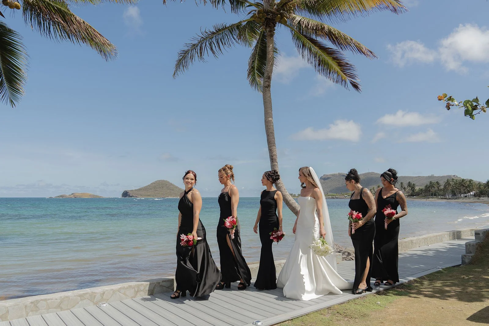 Bride and bridesmaids in black dresses walking along the beach path near a palm tree in St. Lucia