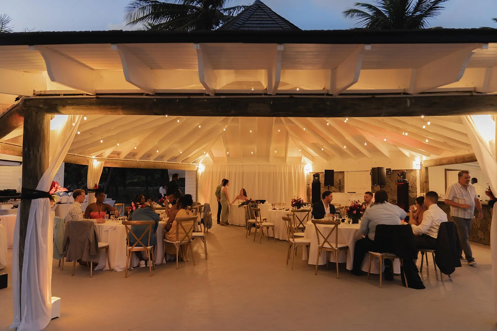 Wide view of the open-air reception pavilion lit up at dusk, wedding guests seated at tables in St. Lucia