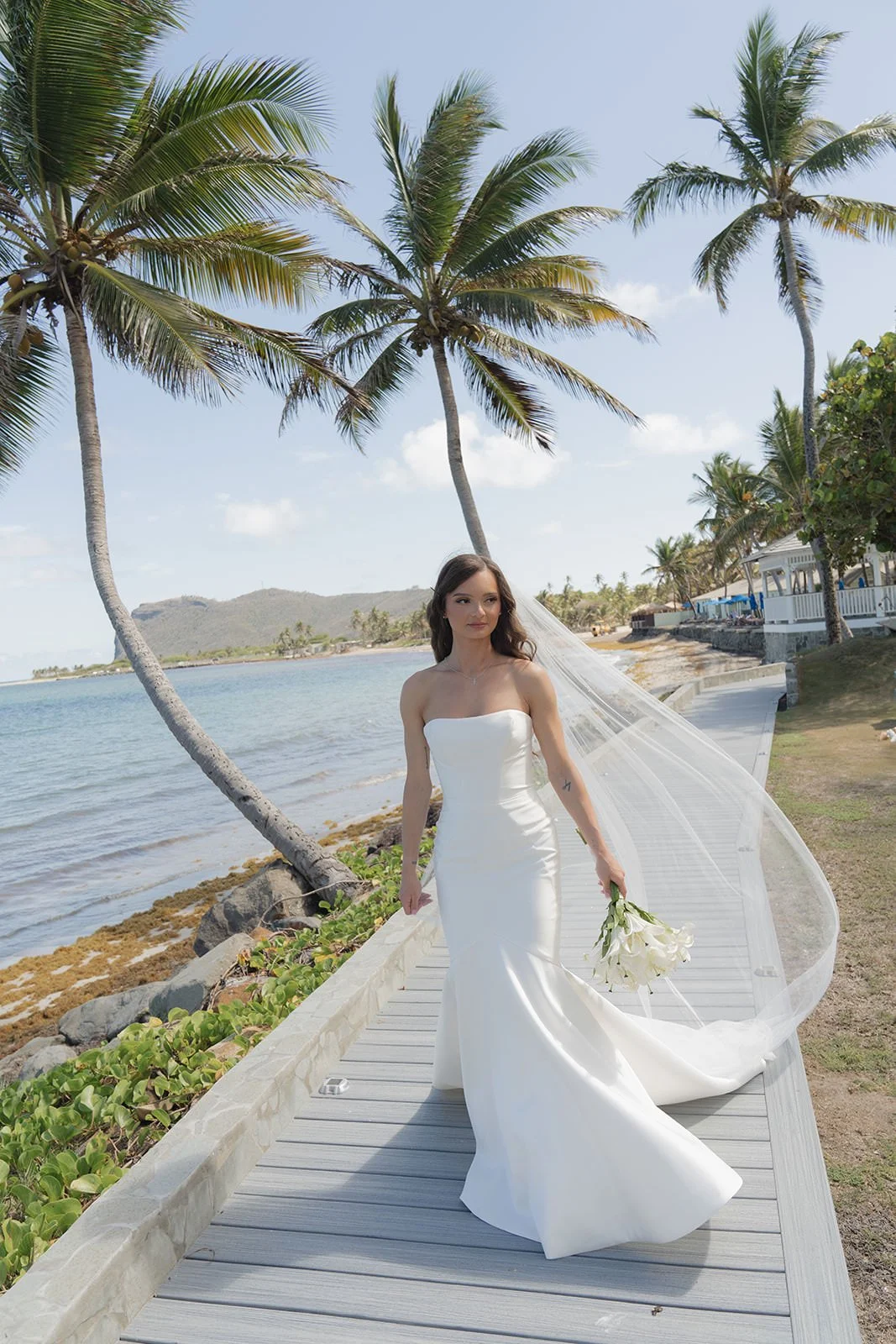 Bride in a strapless gown walking along a St. Lucia beachfront boardwalk, veil trailing in the breeze as she carries her bouquet