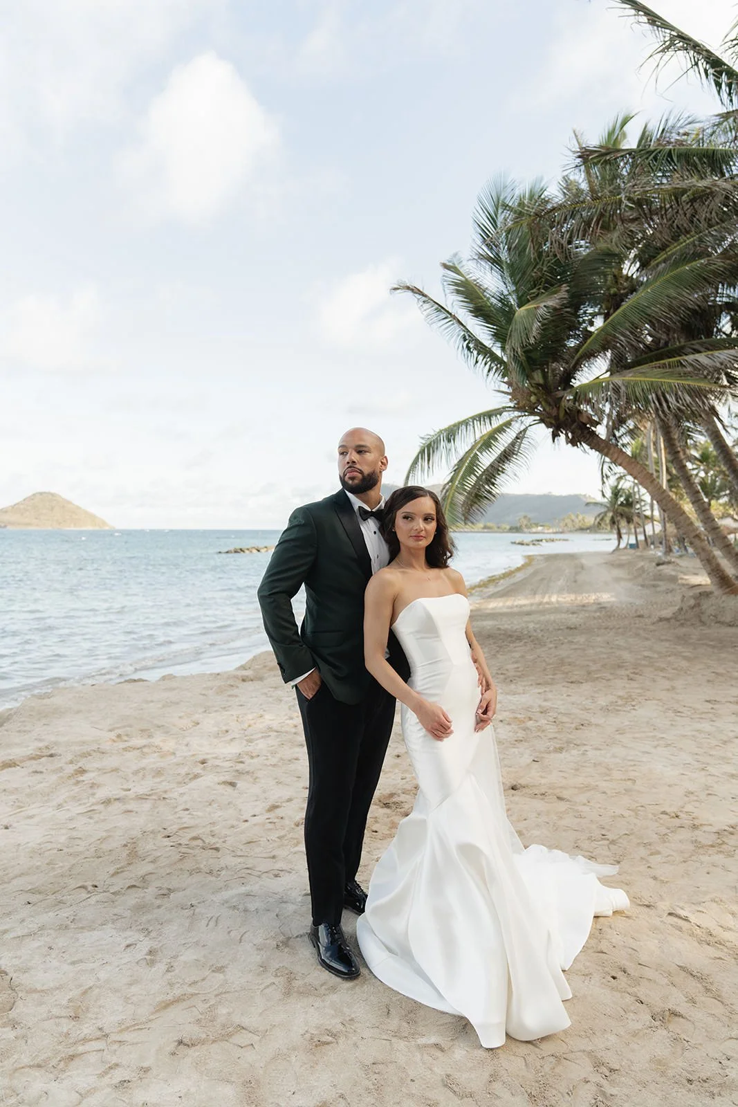Groom in a black tuxedo and bride in a fitted gown posing together on a St. Lucia beach with the ocean and coastline behind them