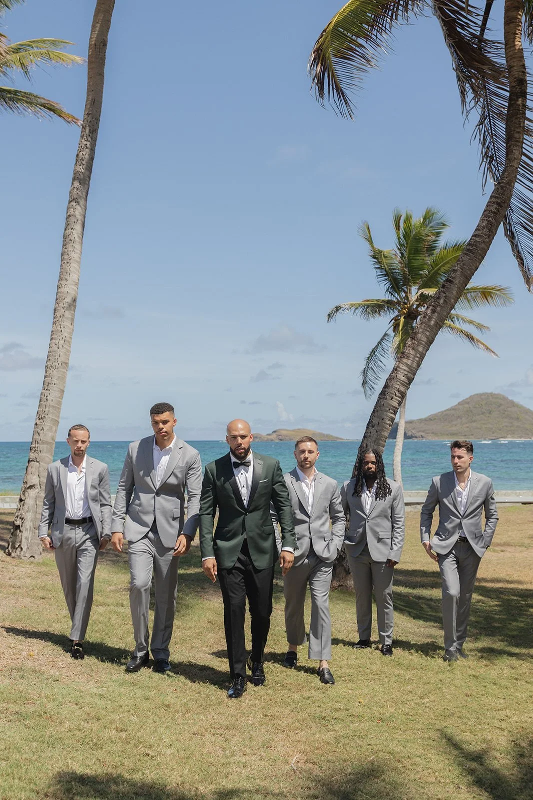 Groom in a dark green suit standing with groomsmen in matching gray suits on a St. Lucia beach, palm trees and hills in the background