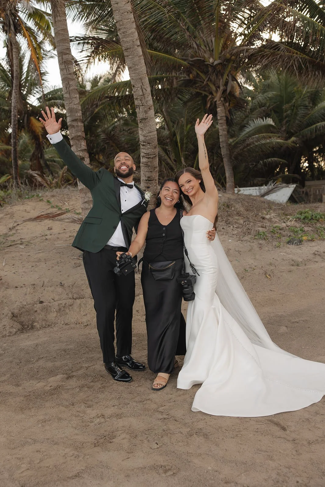 Bride, groom, and a bridesmaid raising their hands and celebrating together on a St. Lucia beach lined with palm trees