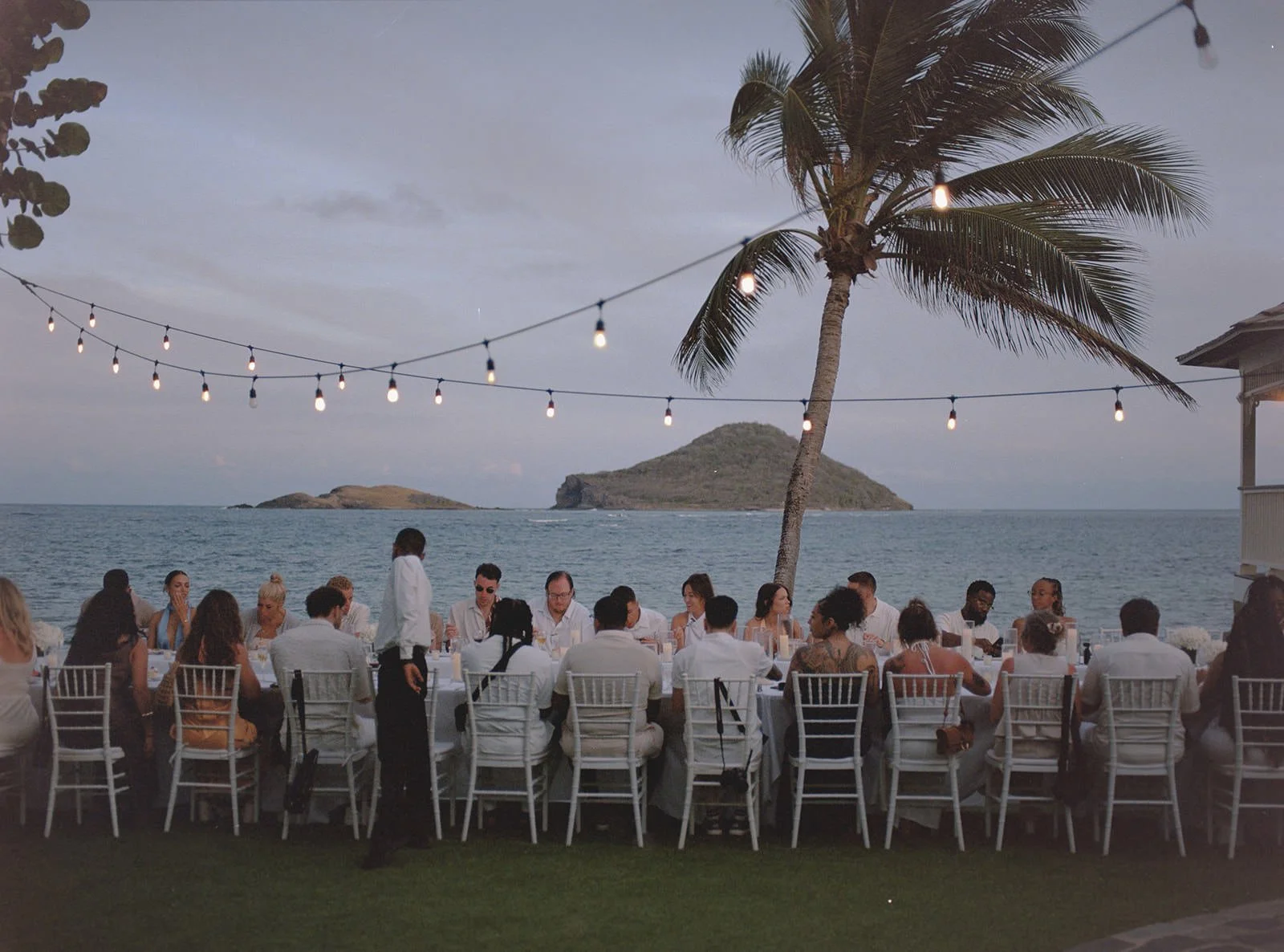 Wedding guests seated at a long dinner table strung with lights, island and ocean in the background, St. Lucia