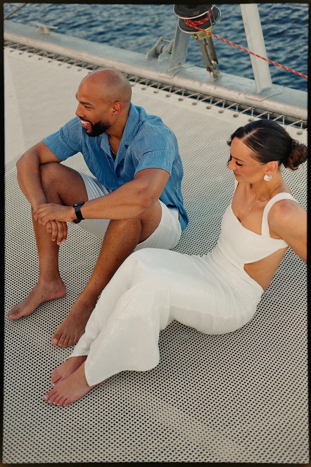 Groom and bride sitting together on the catamaran's trampoline netting, laughing and talking during the wedding weekend sailing excursion