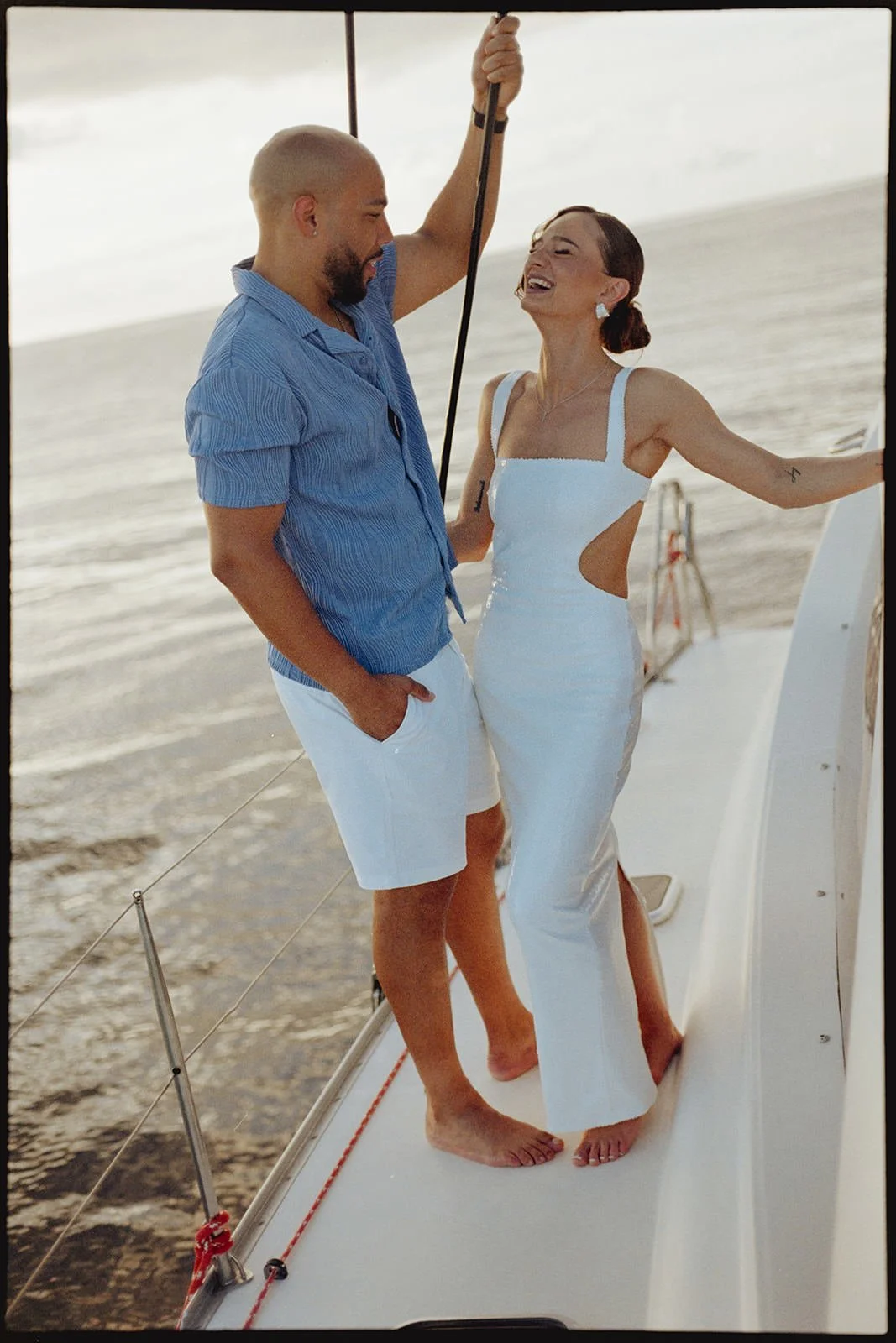 Groom holding onto the catamaran's rigging as the bride leans back playfully in her white dress, both barefoot on the bow