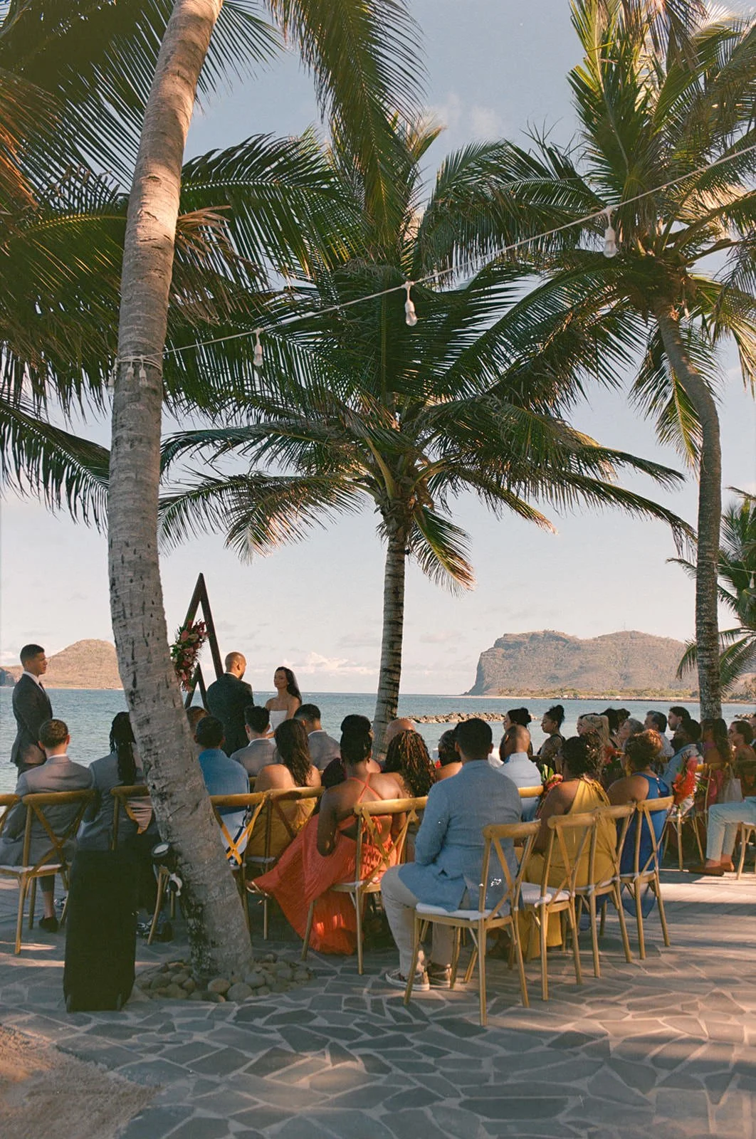 Wedding guests seated for the reception at sunset beneath palm trees, ocean and mountains in the background in St. Lucia