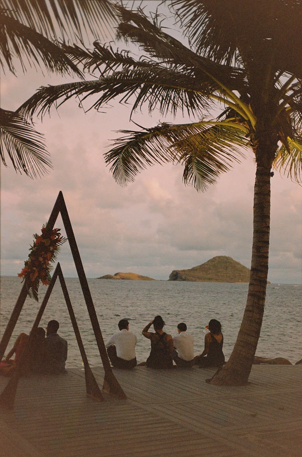 Sepia-toned view of wedding guests seated near the triangular ceremony arch, palm trees and ocean in the background in St. Lucia