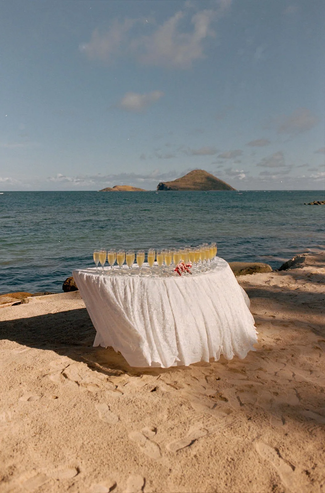 Small sweetheart table with a white linen cloth and drink glasses set up on the sand, ocean and small island in the background at the St. Lucia wedding