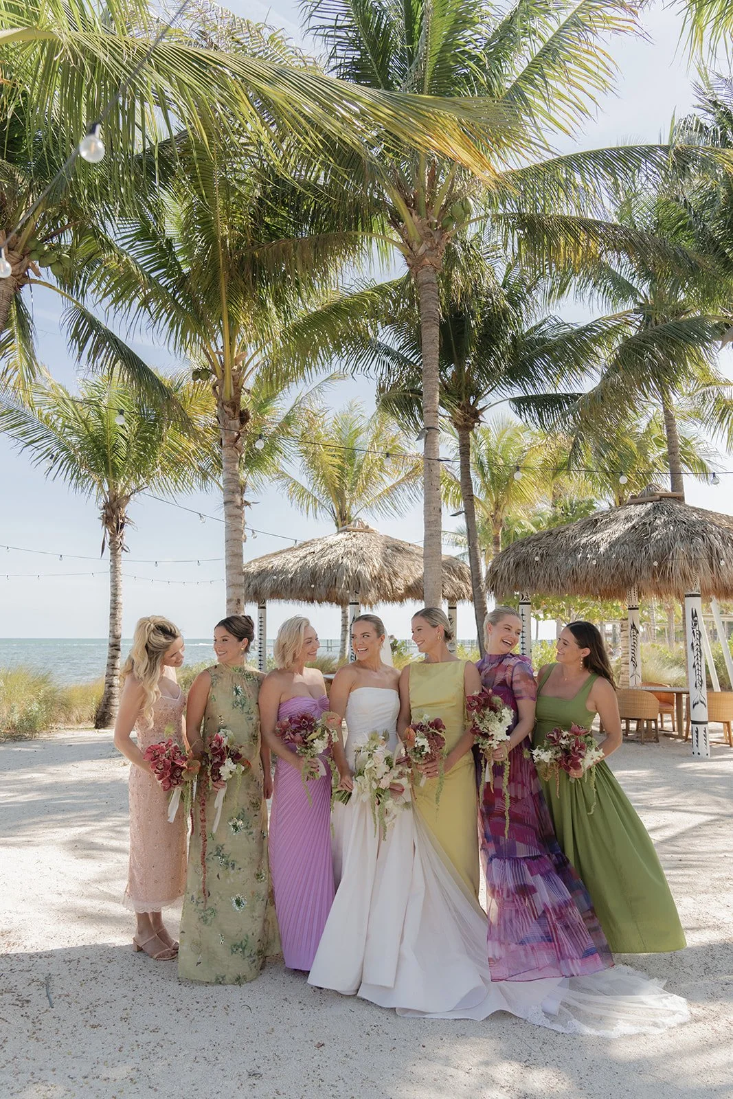 Bride and bridesmaids in mismatched pastel gowns pose among palm trees on the beach at the Islander Resort in Islamorada, Florida Keys