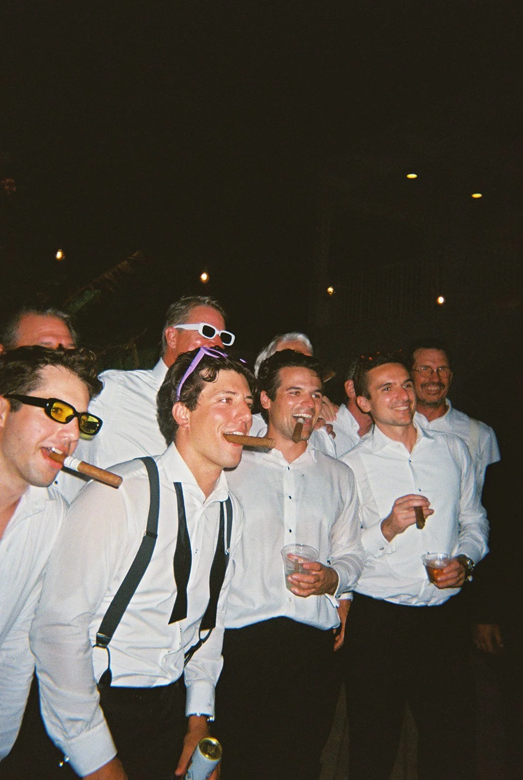 Groomsmen in white shirts and suspenders smoking cigars and laughing at night during this Islamorada, Florida Keys destination wedding