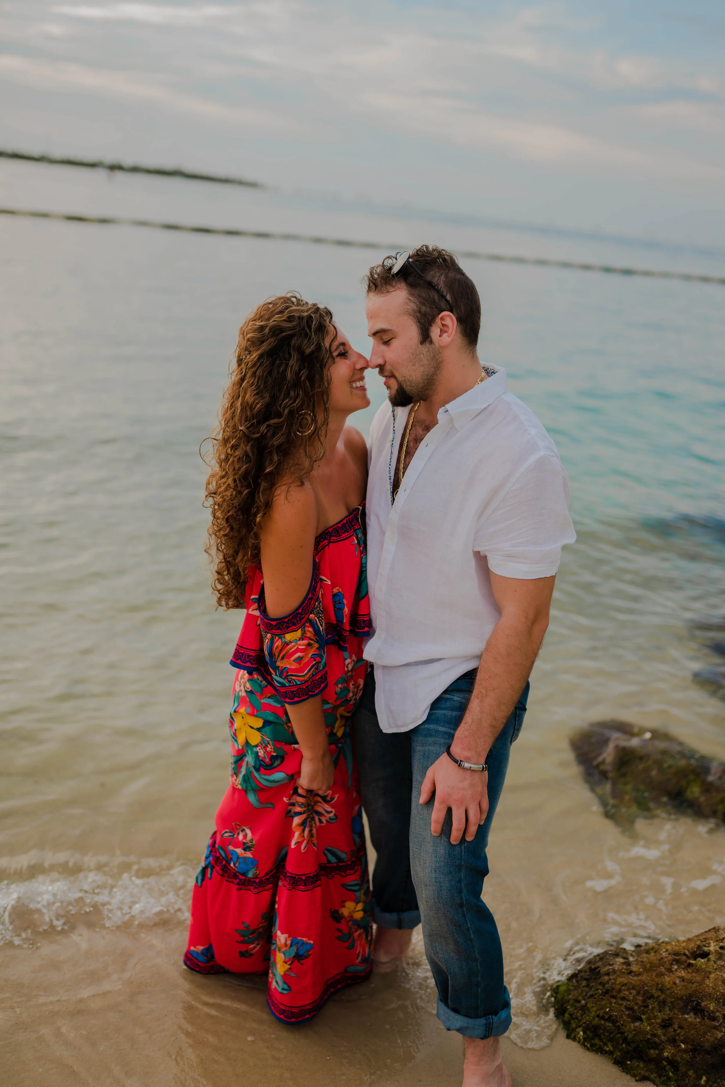 A couple standing close together on a beach, with their foreheads touching, smiling, and looking into each other's eyes.