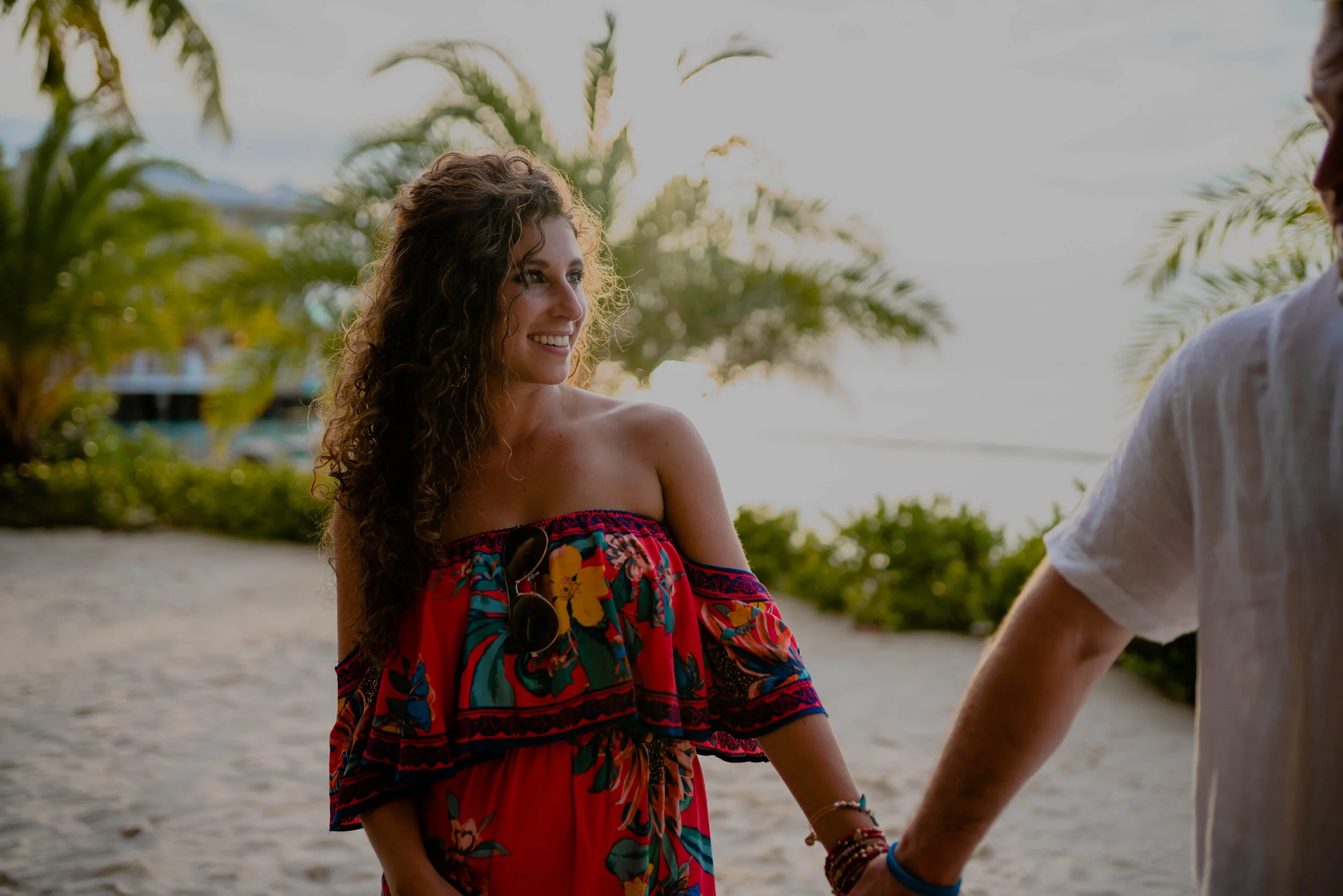 A woman smiling and holding hands with a man on a beach with palm trees, sand, and the ocean in the background during sunset.