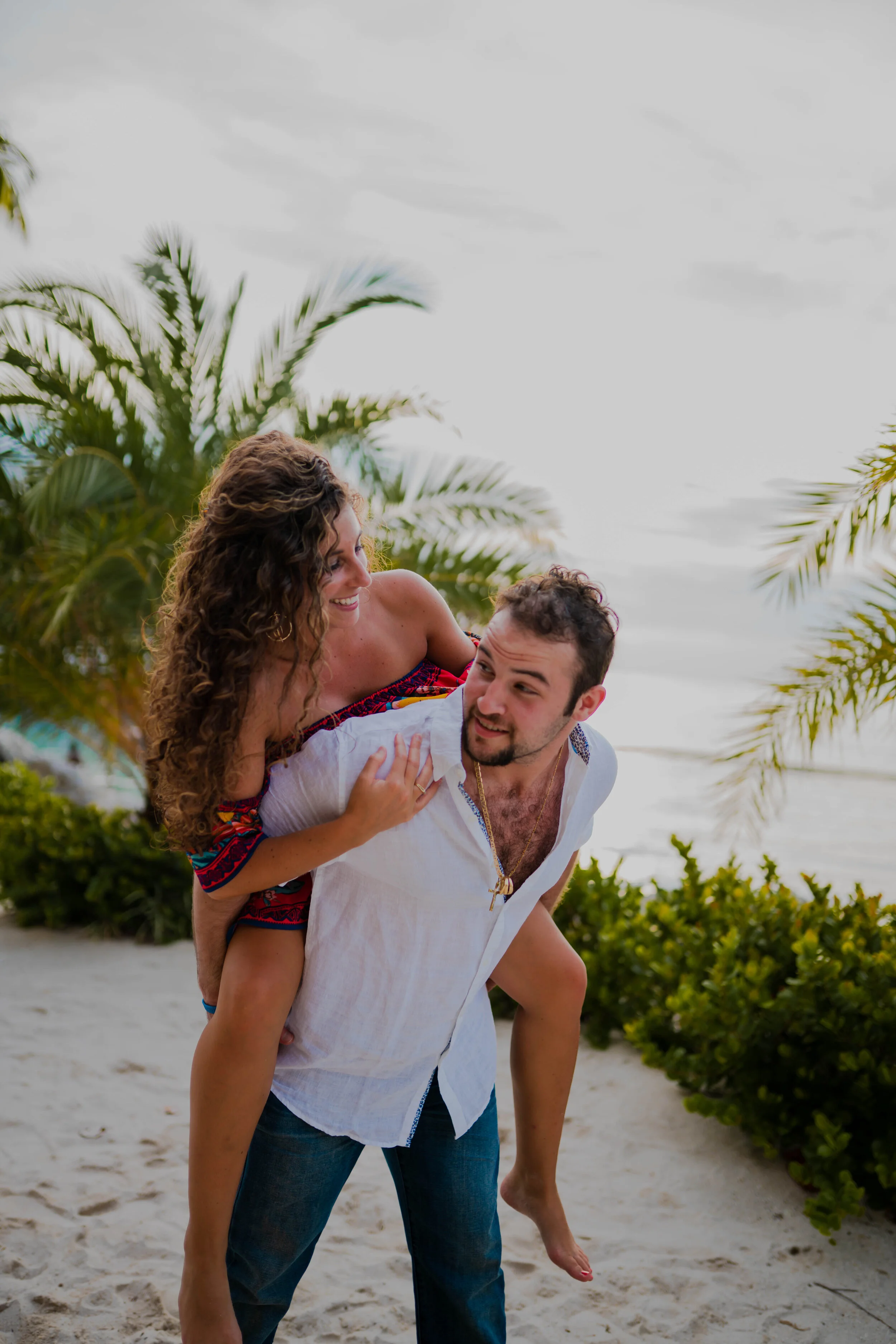 A man giving a woman a piggyback ride on a beach with palm trees and water in the background, both smiling and enjoying themselves.