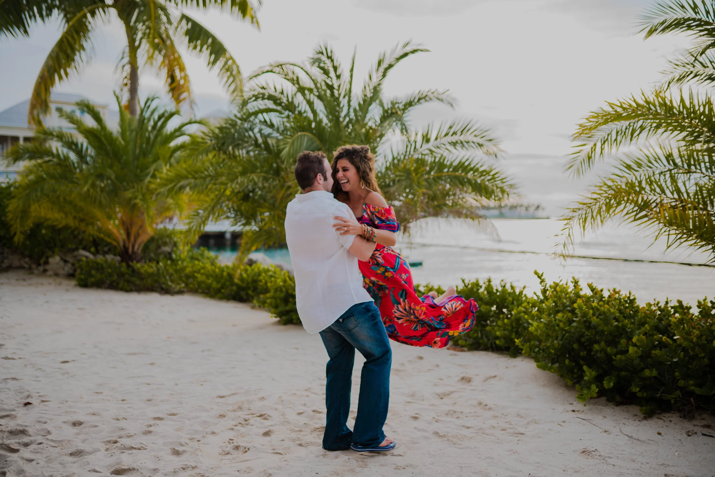 A man and woman are dancing and laughing on a beach with palm trees in the background.