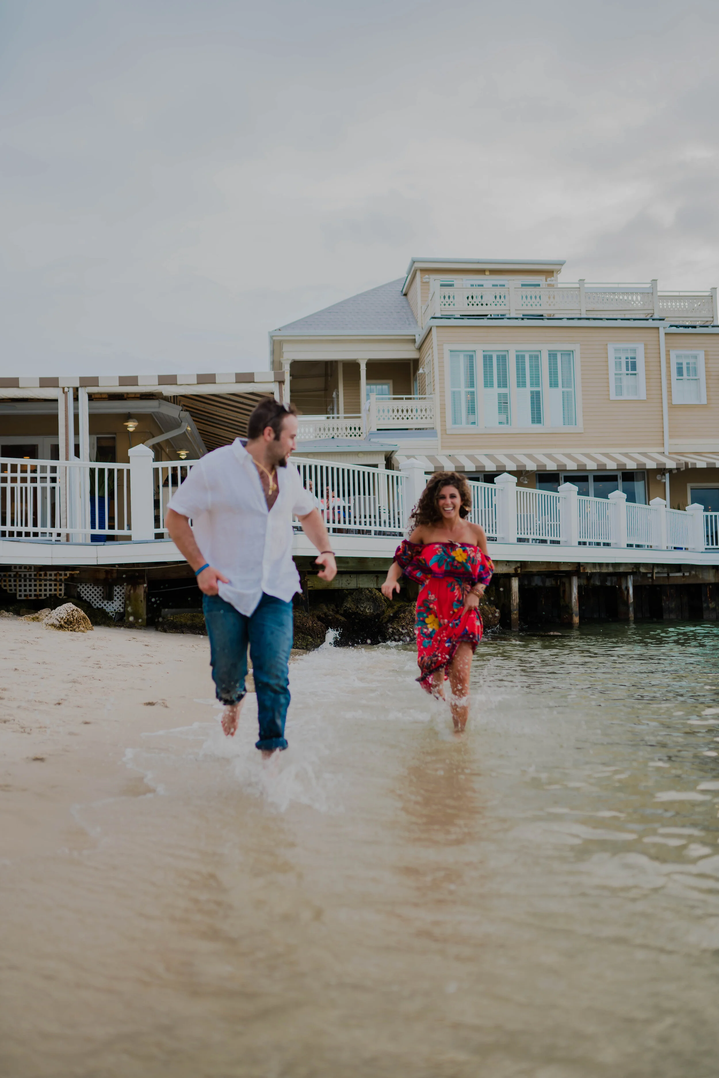 A man and woman running in shallow water at the beach near a large beach house, both smiling and enjoying the moment.