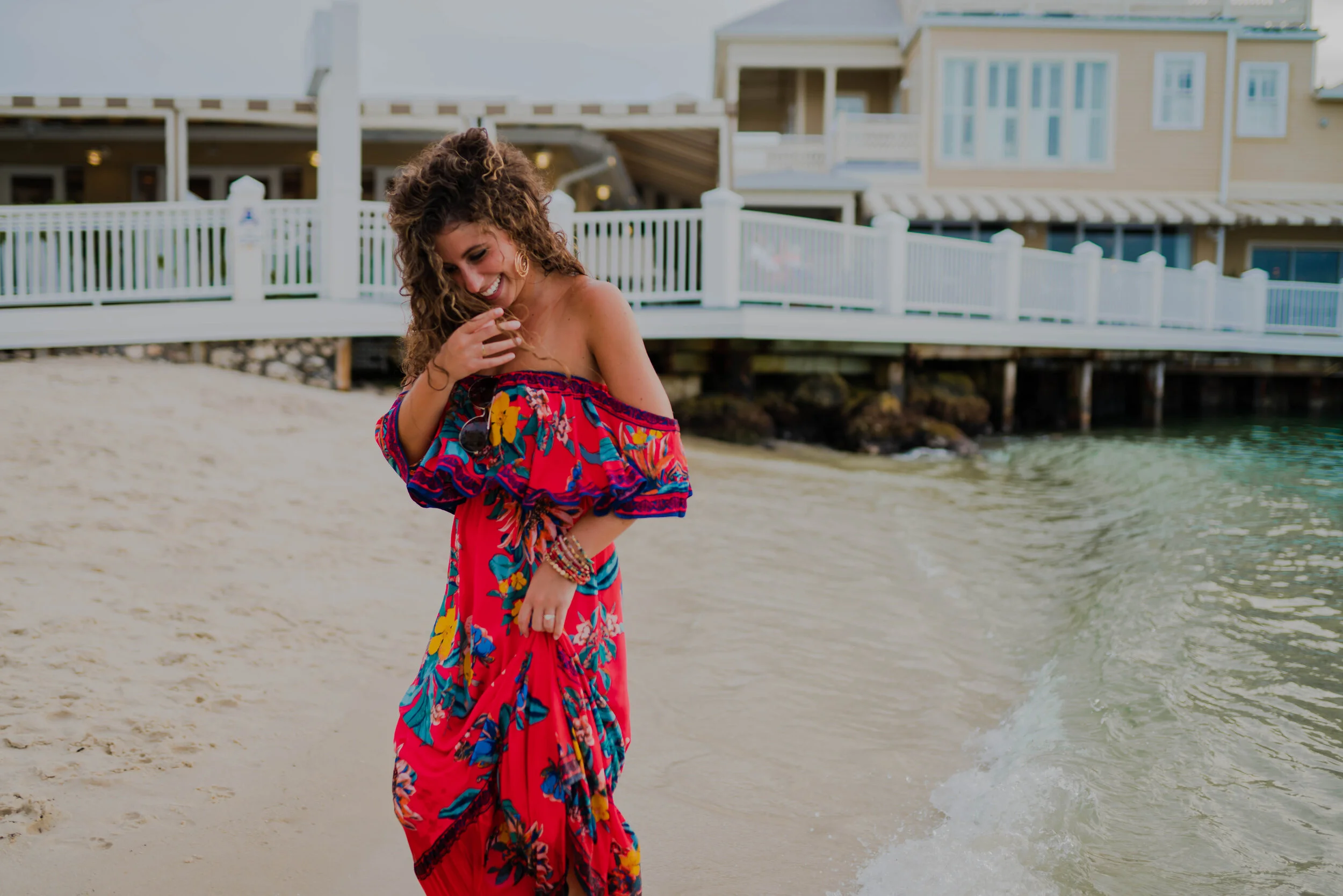 Woman in a colorful off-the-shoulder dress smiling and holding her dress on a sandy beach near water, with houses and a pier in the background.
