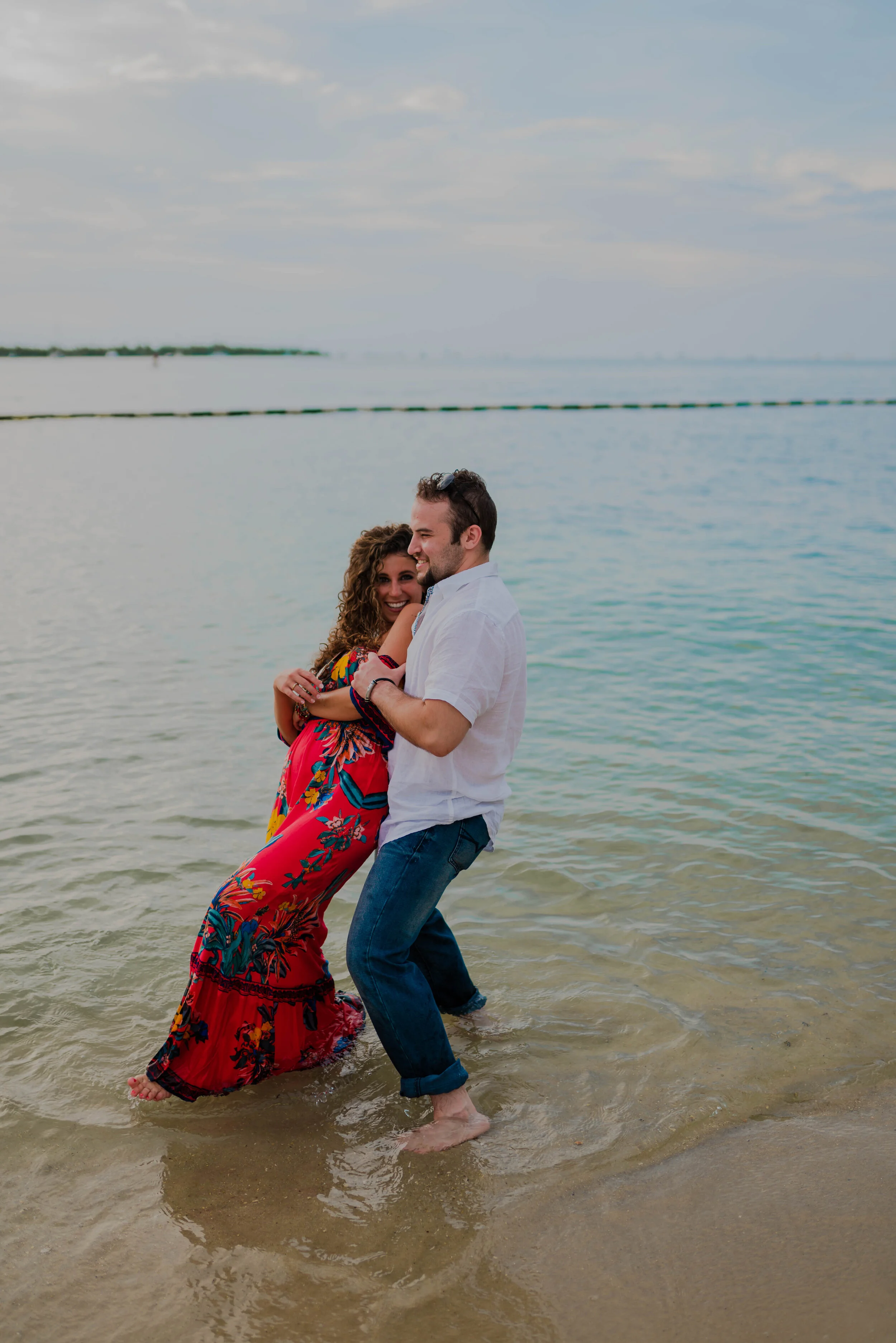 A couple standing in shallow ocean water, smiling and embracing, with a cloudy sky and a distant shoreline in the background.