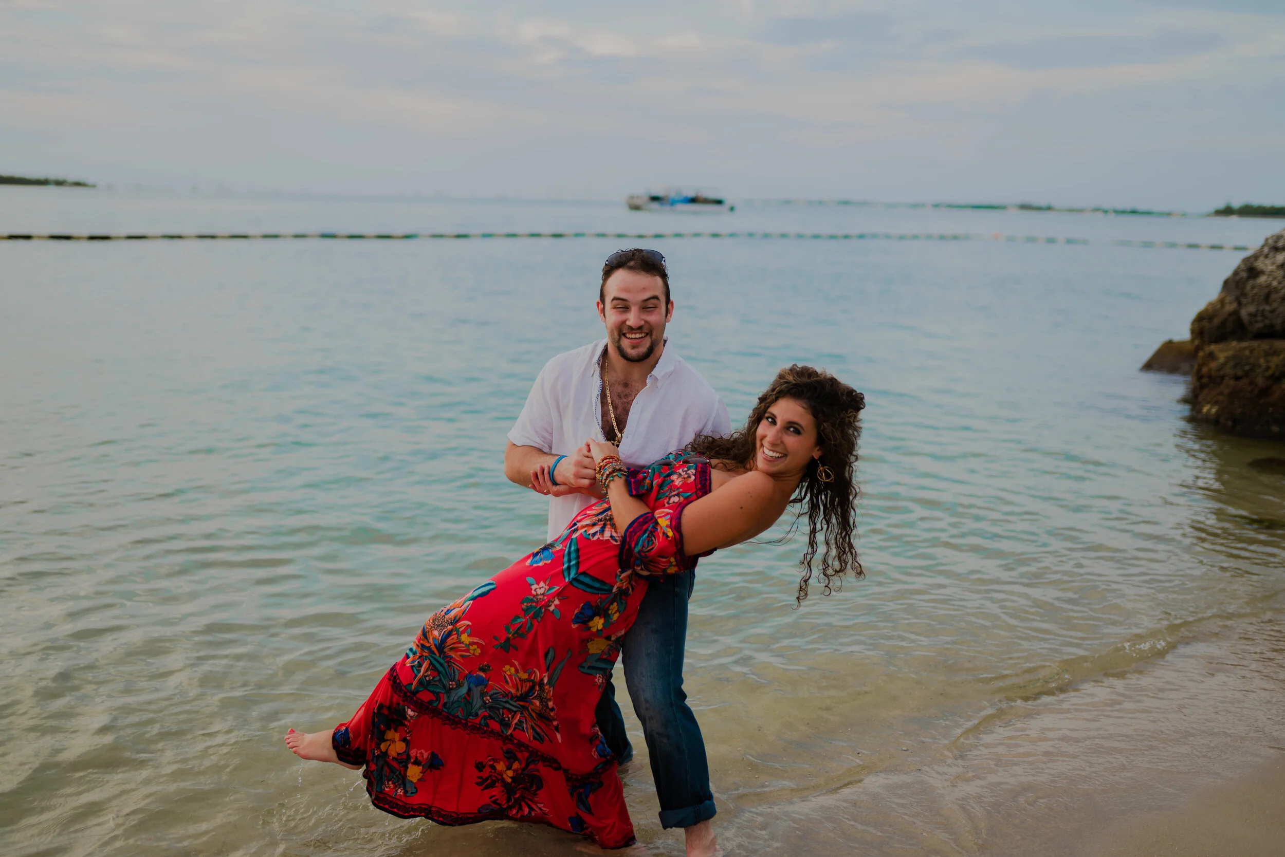 A man and woman smiling and posing in the shallow waters of a beach, with the man dipping the woman playfully as they enjoy a sunny day near the ocean.