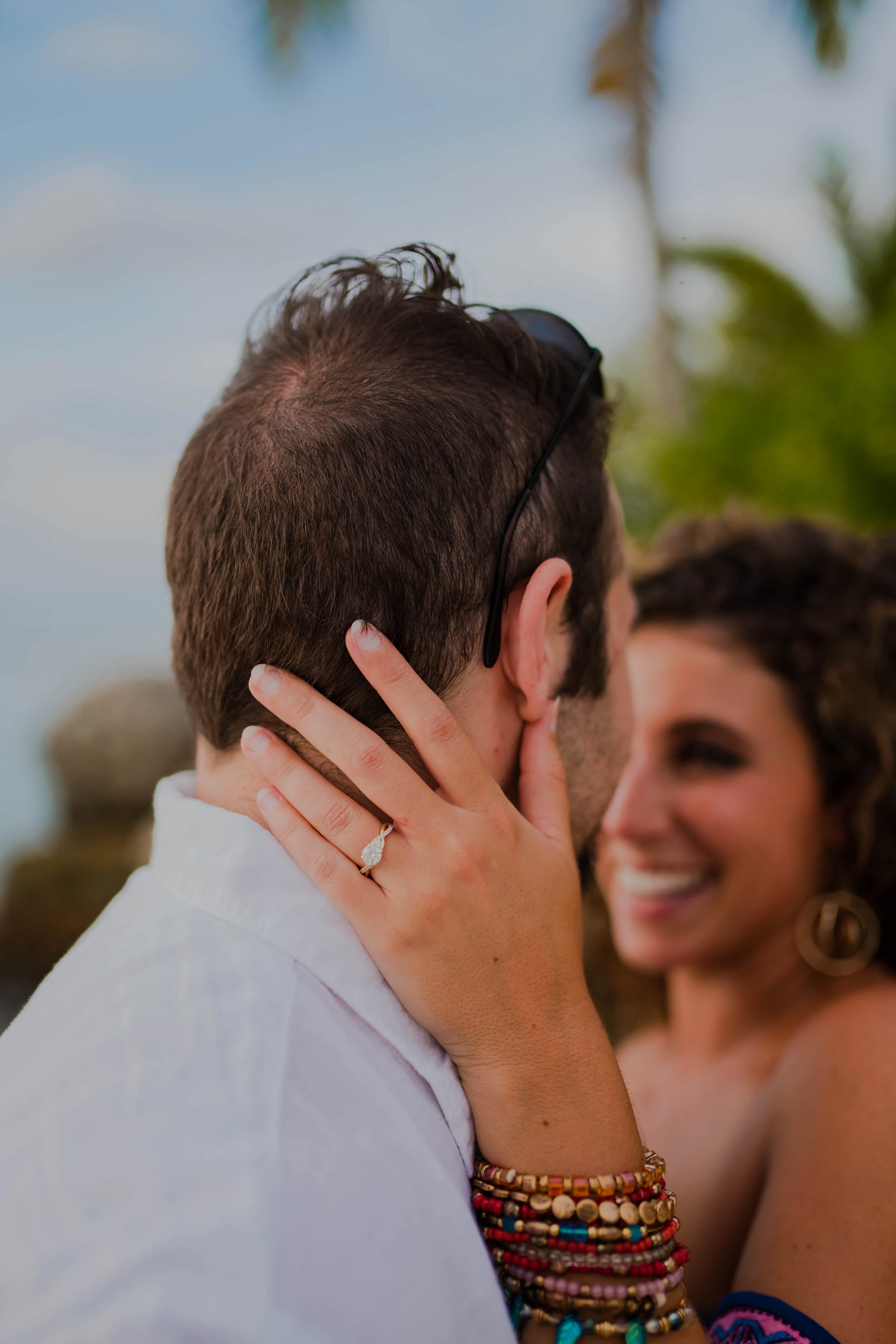 A couple kissing outdoors, the woman wearing multiple beaded bracelets and an engagement ring, with palm trees in the background.