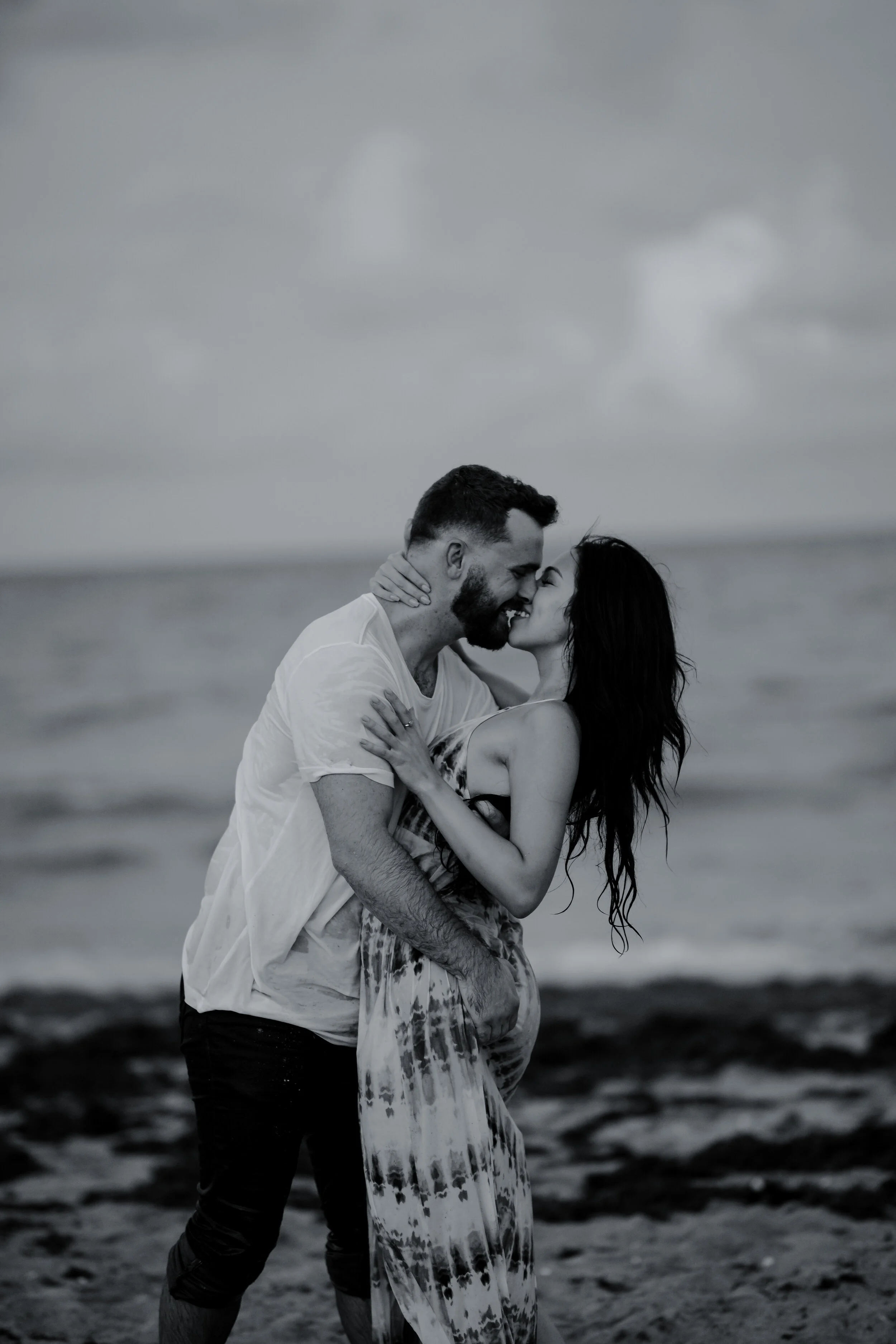 A couple embracing and smiling at each other on the beach, with the ocean and cloudy sky in the background, in black and white.
