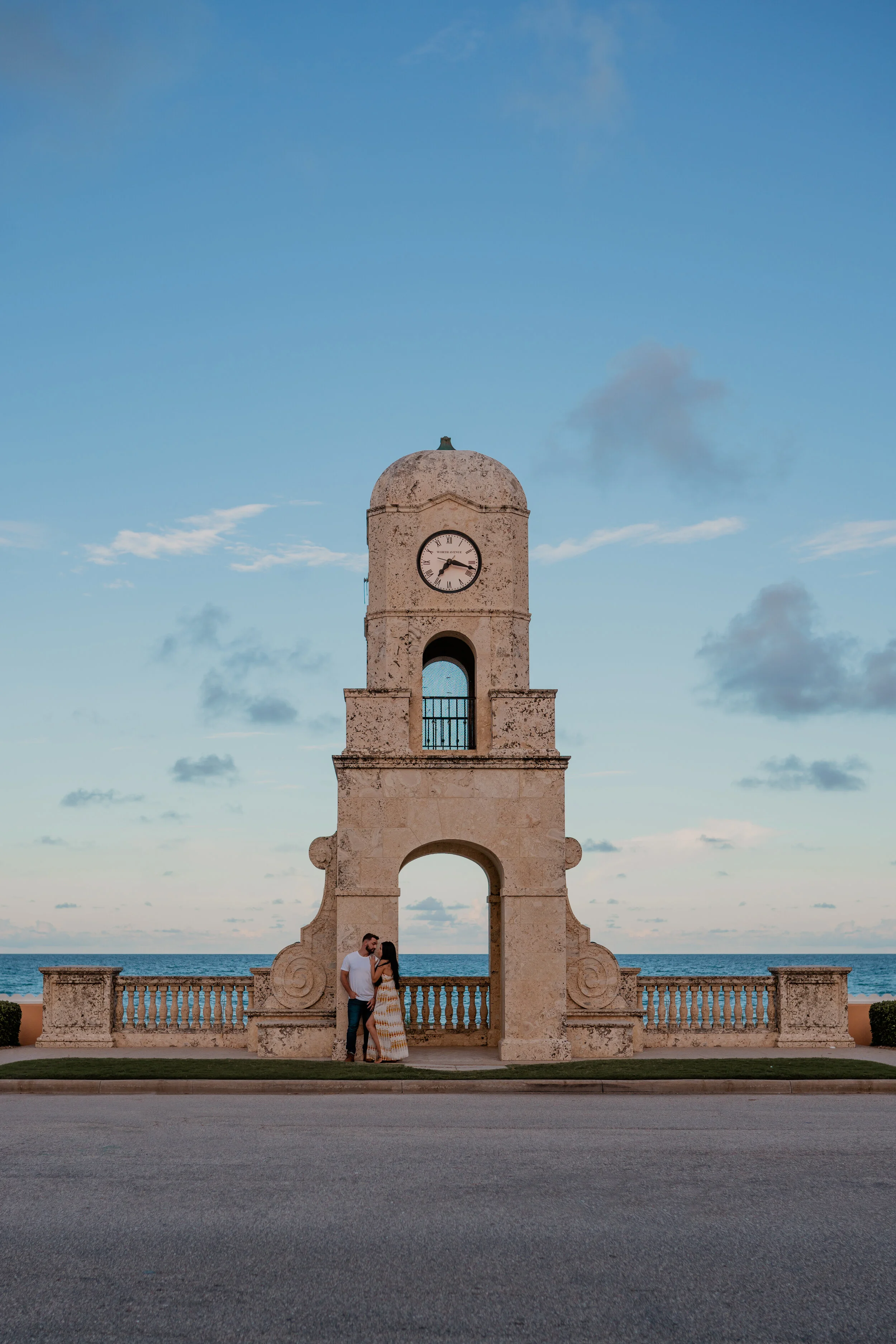 A man and woman standing close together, kissing under an old stone clock tower near the ocean during sunset.