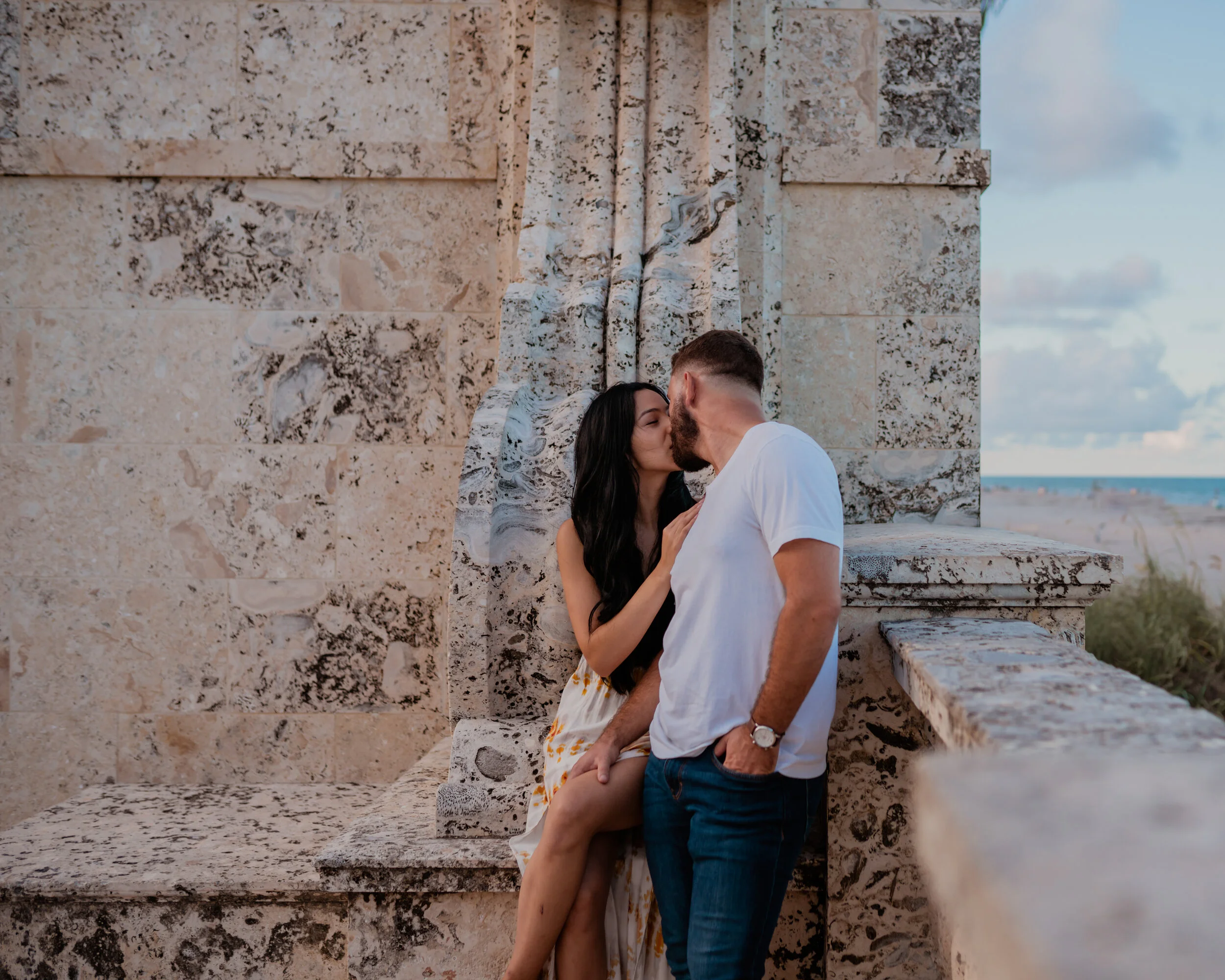 A couple kissing near a stone structure on the beach during sunset.
