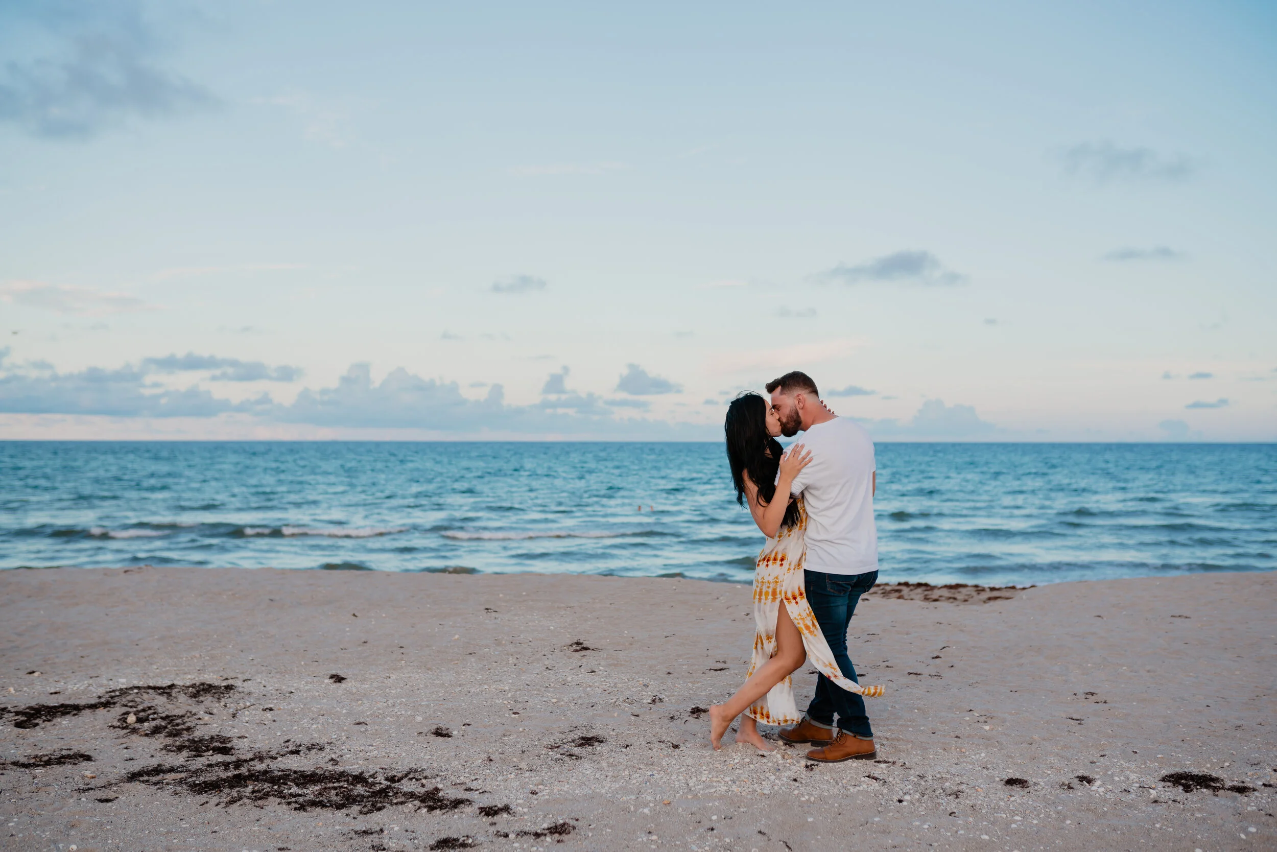 A couple sharing a kiss on a beach during sunset, with ocean and sky in the background.