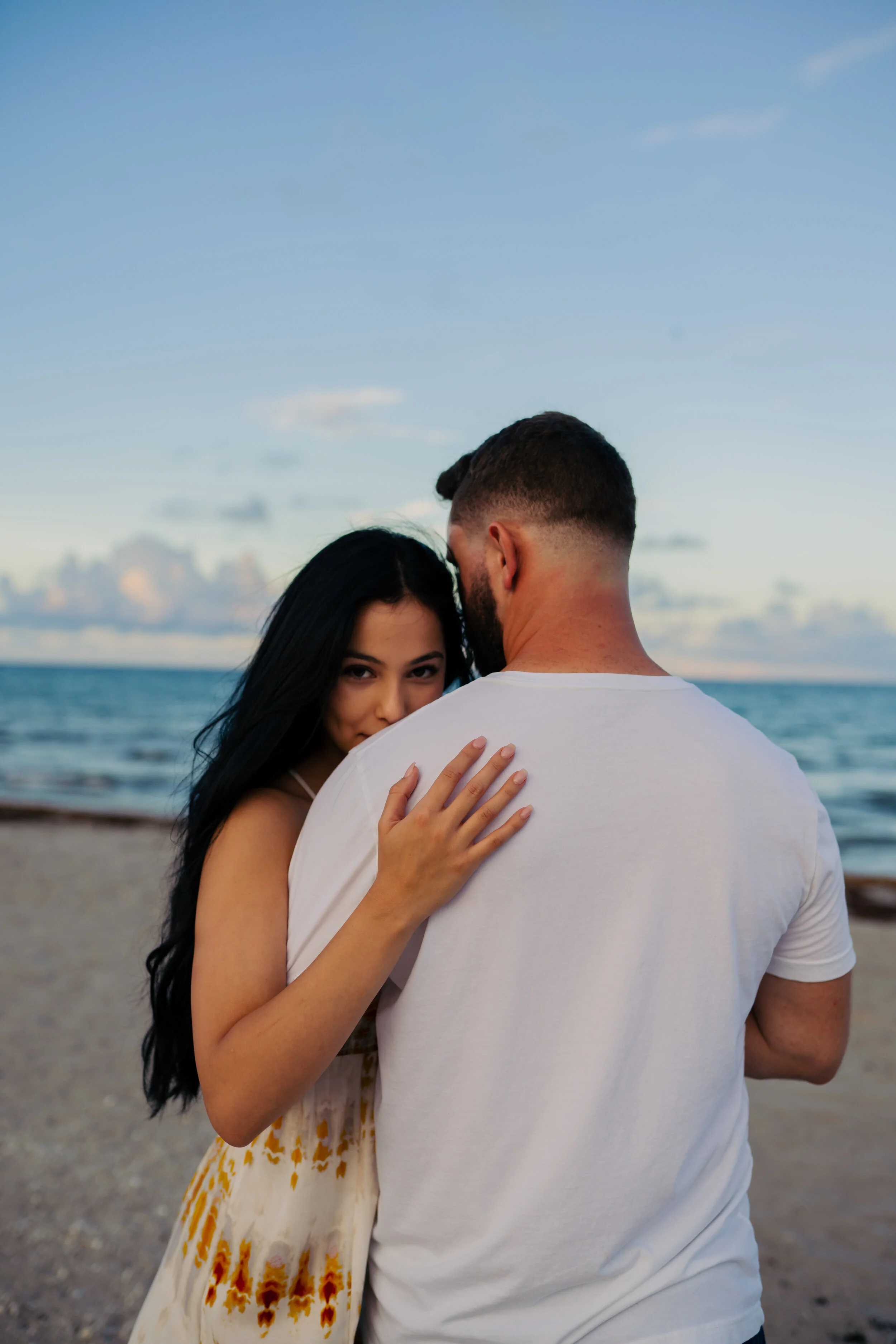 A woman with long dark hair and wearing a light-colored dress clinging to a man with a beard and short hair, wearing a white t-shirt, on a beach at sunset, with the ocean and sky in the background.