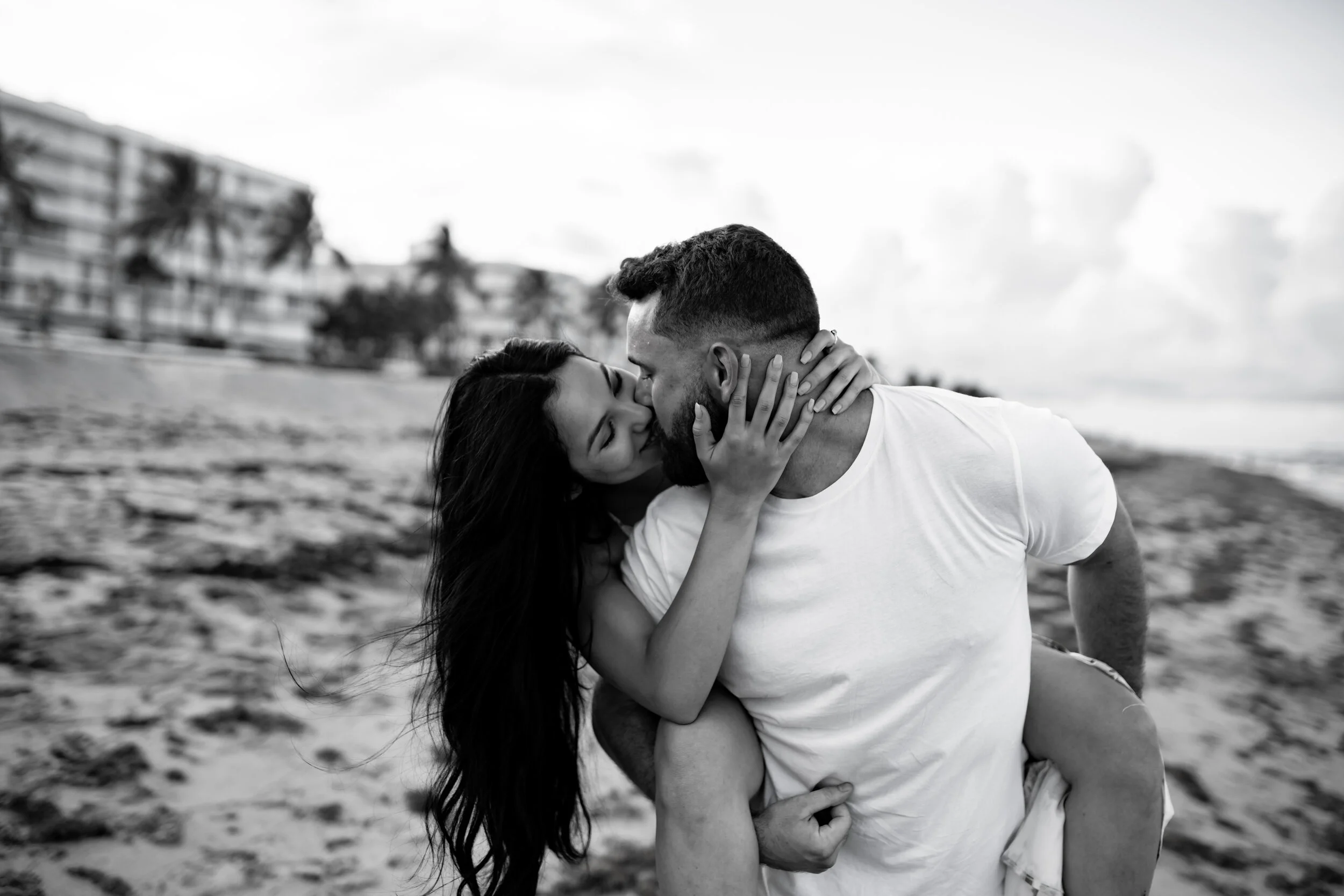 A couple kissing on the beach, with the man giving the woman a piggyback ride, in black and white.