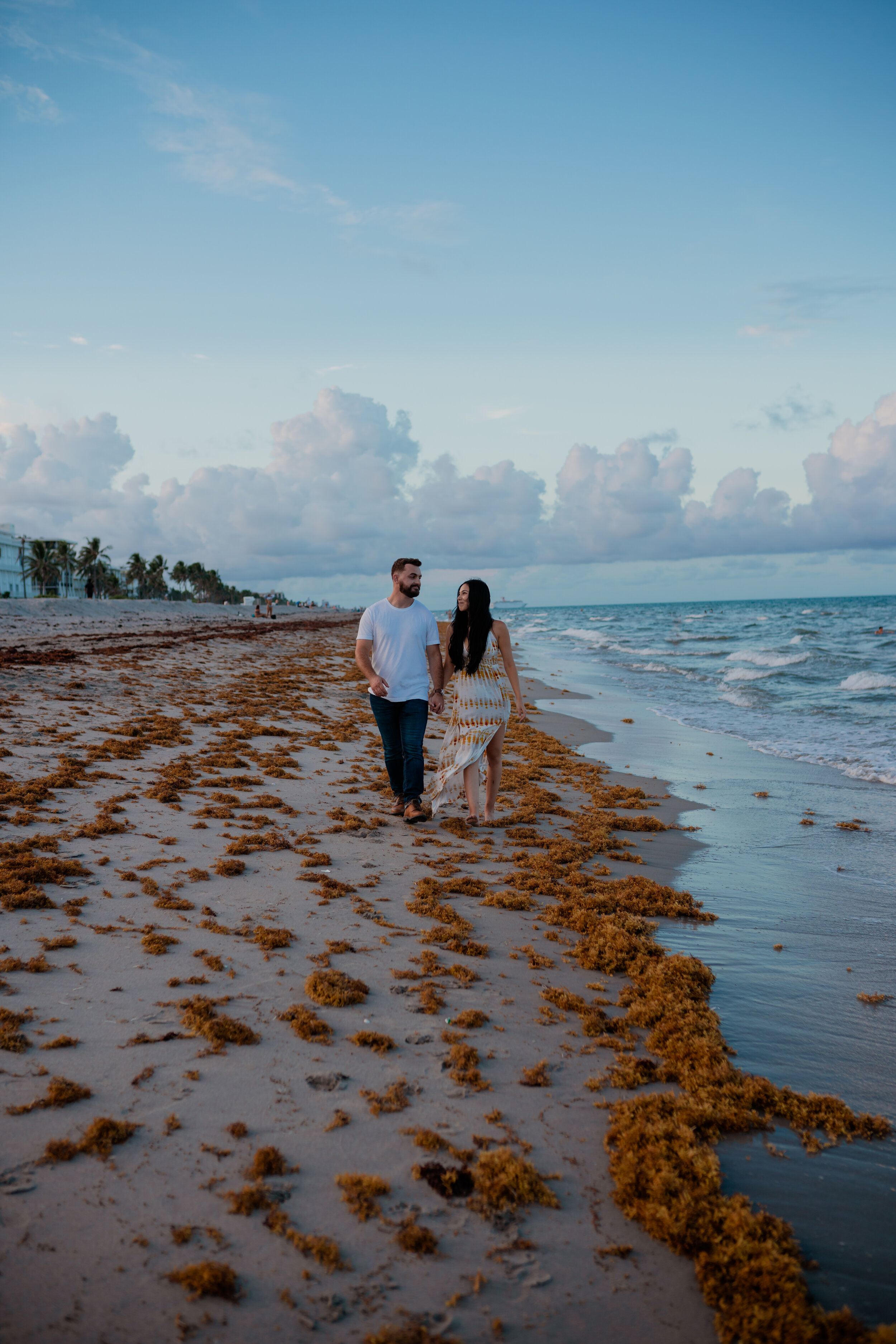 A couple walks hand in hand along a sandy beach with seaweed, with a partly cloudy sky and ocean in the background.