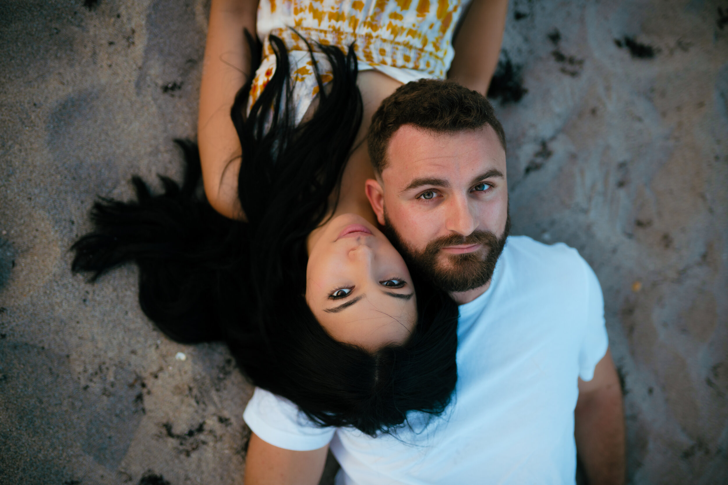 A man and woman lying on the sand at the beach, with the woman’s head resting on the man’s shoulder, both looking at the camera.