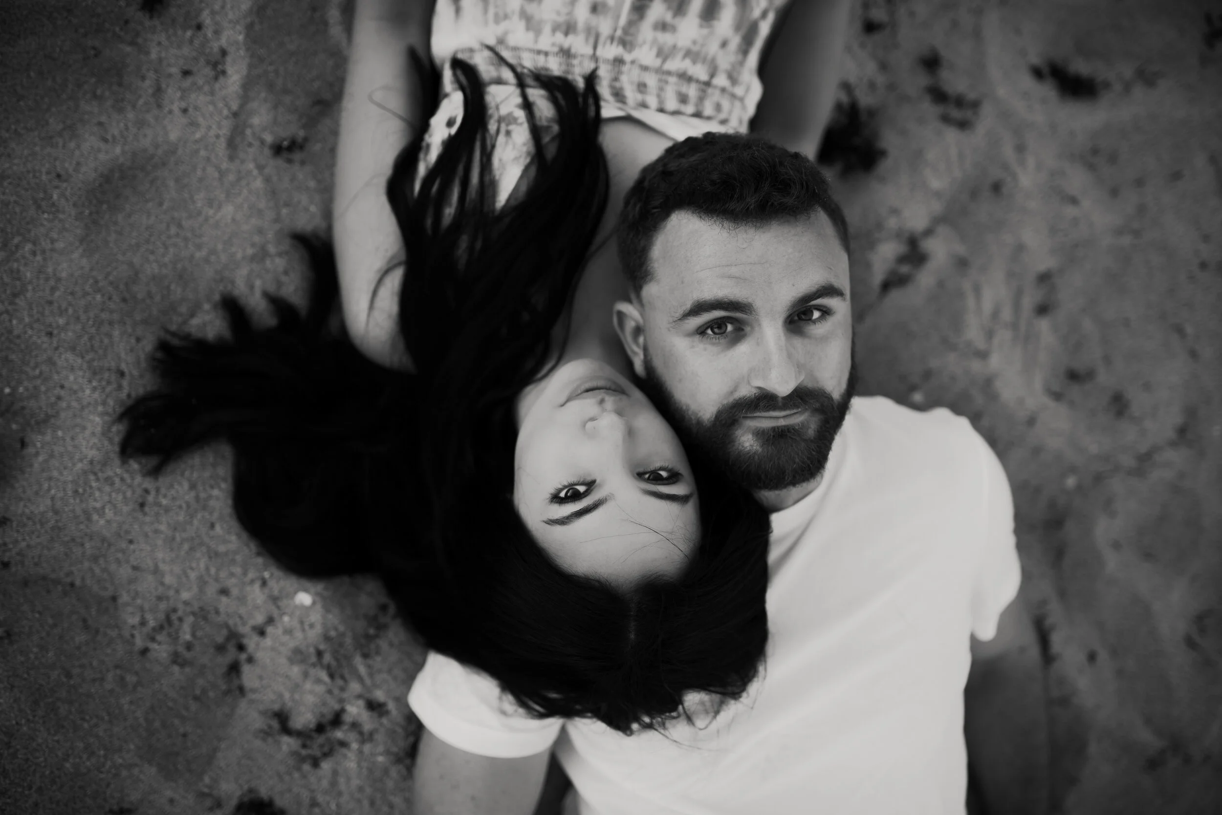 A black and white photo of a man and a woman lying on the ground, with the woman resting her head on the man's chest, both looking upward towards the camera.