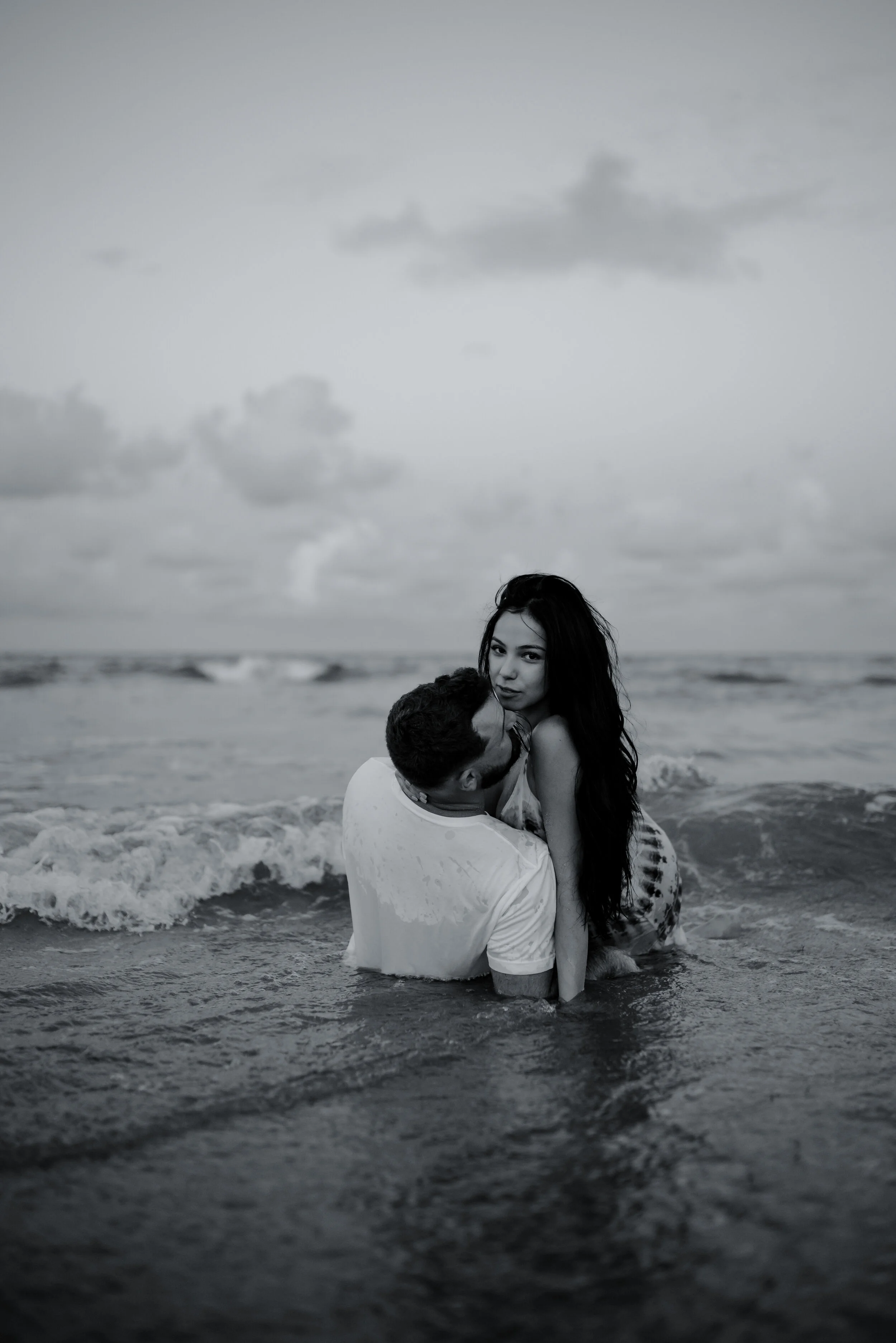 A black-and-white photo of a man holding a woman in the ocean, with waves around them and cloudy sky above.