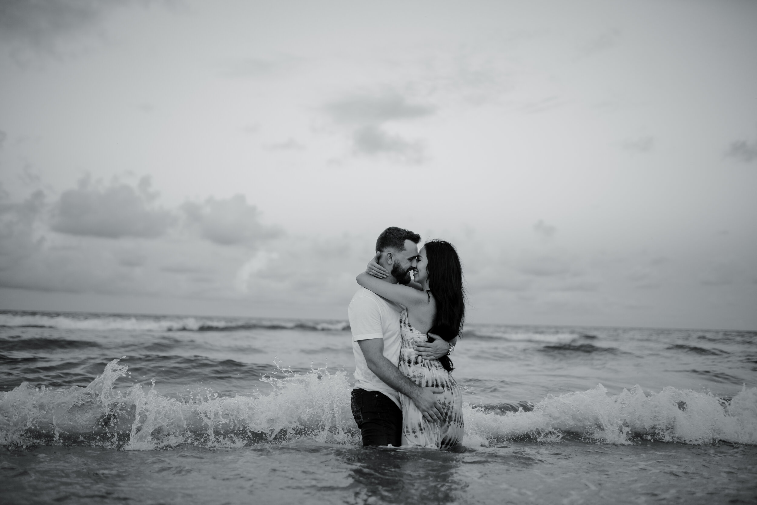 A couple embracing and smiling at each other in the ocean with waves around them, under a cloudy sky.