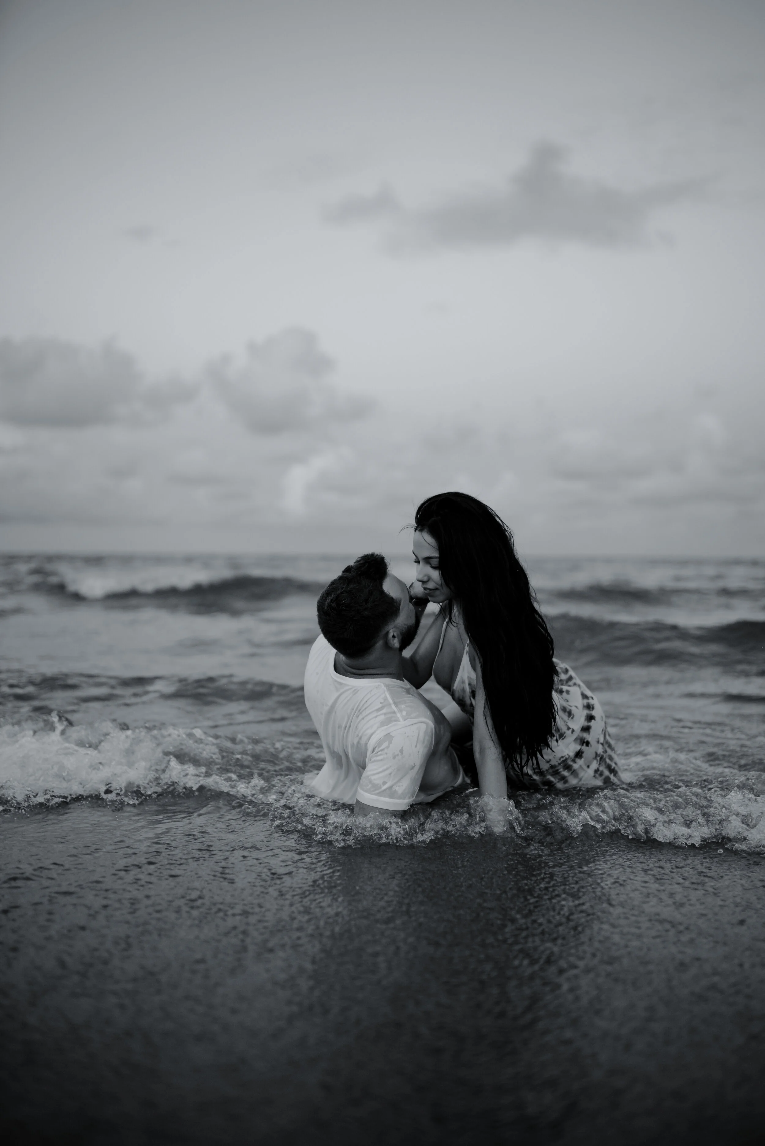 A couple in the ocean, embracing and touching noses, with cloudy sky above.