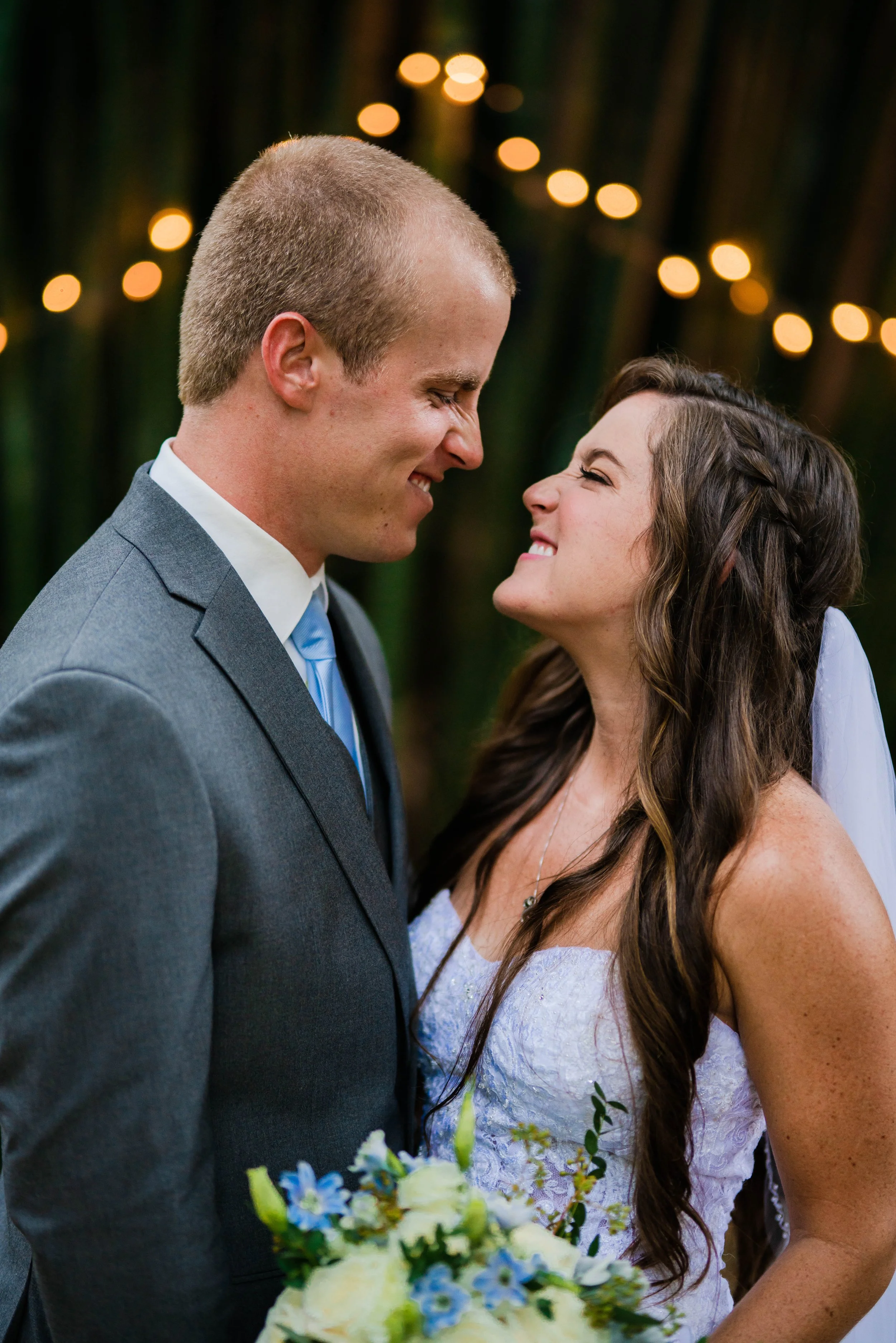 A happy bride and groom close together, standing outdoors with blurred string lights in the background.