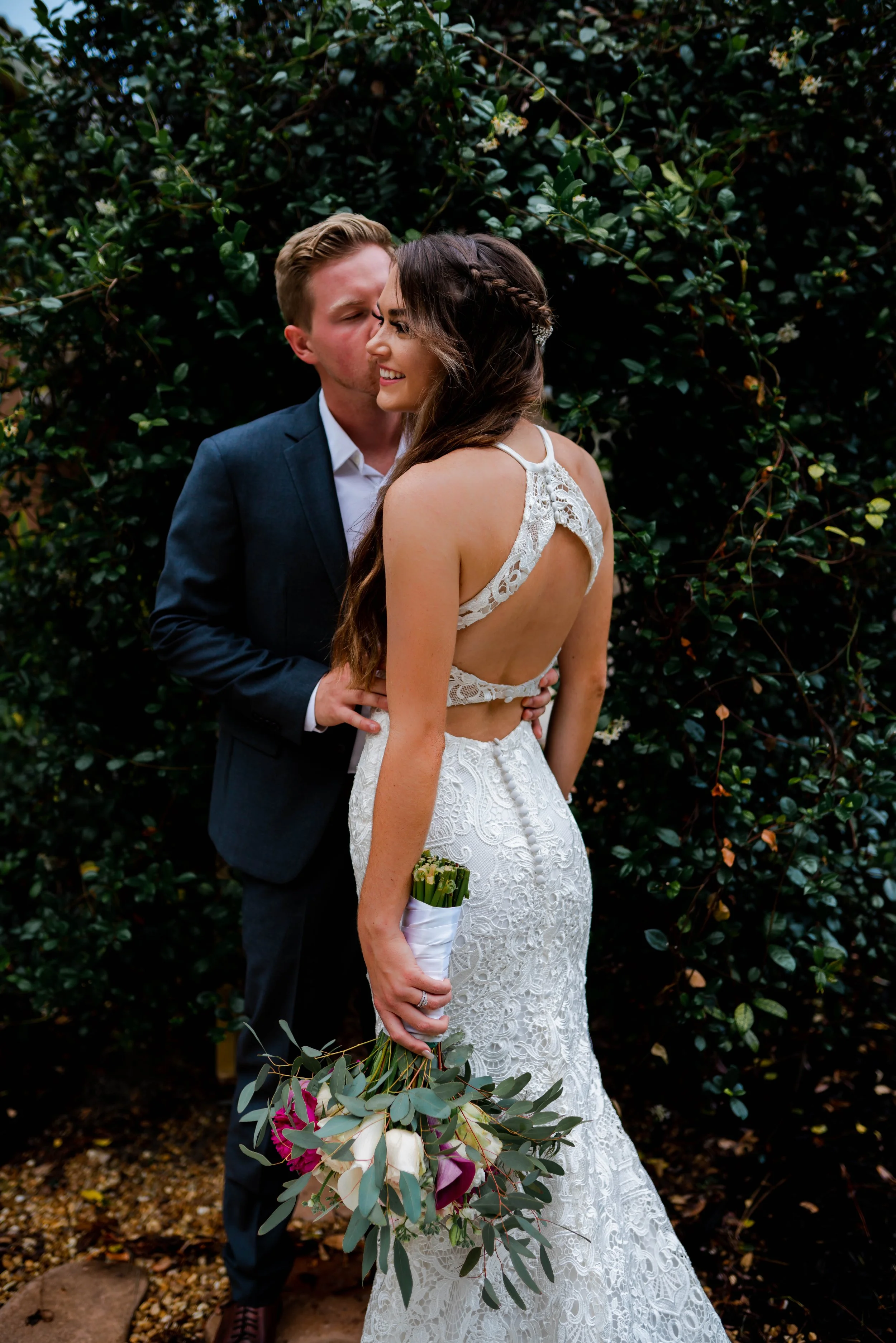A bride in a white lace wedding gown holding a bouquet of flowers, and a groom in a dark suit, sharing a kiss in front of green foliage.