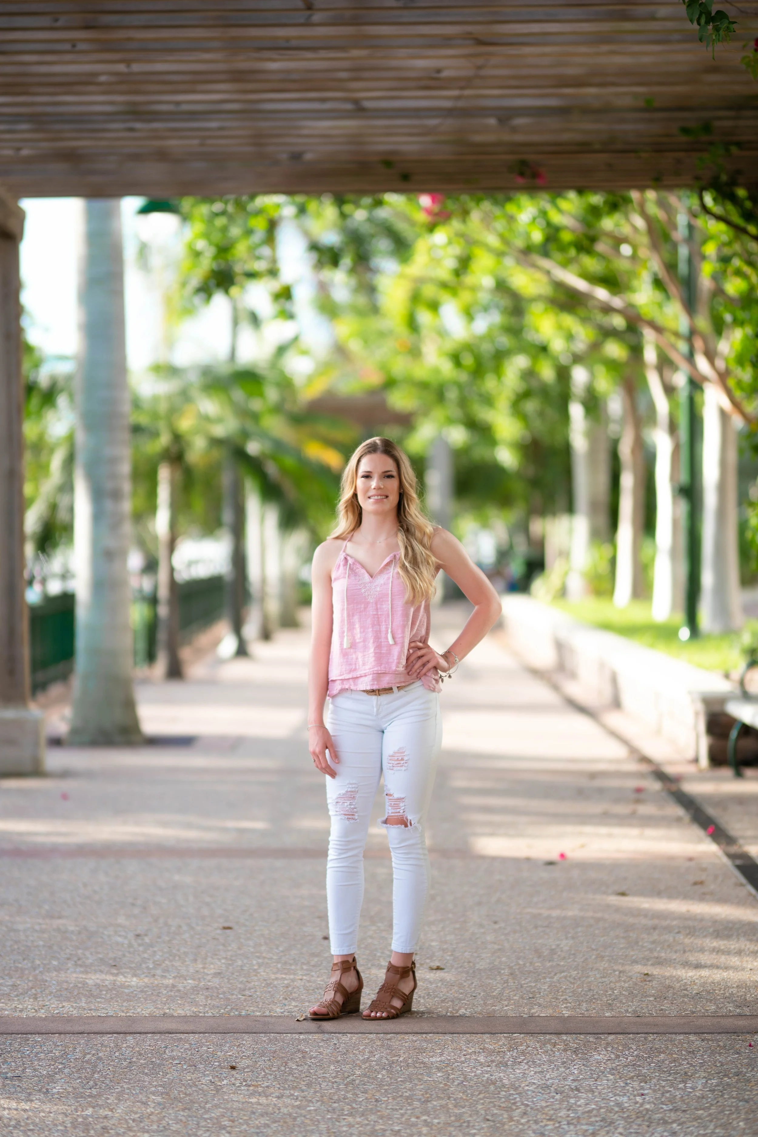 A woman with long, wavy blonde hair stands on a sunny sidewalk in a park, wearing a pink sleeveless top, white ripped jeans, and brown heeled sandals, smiling at the camera with trees and greenery in the background.