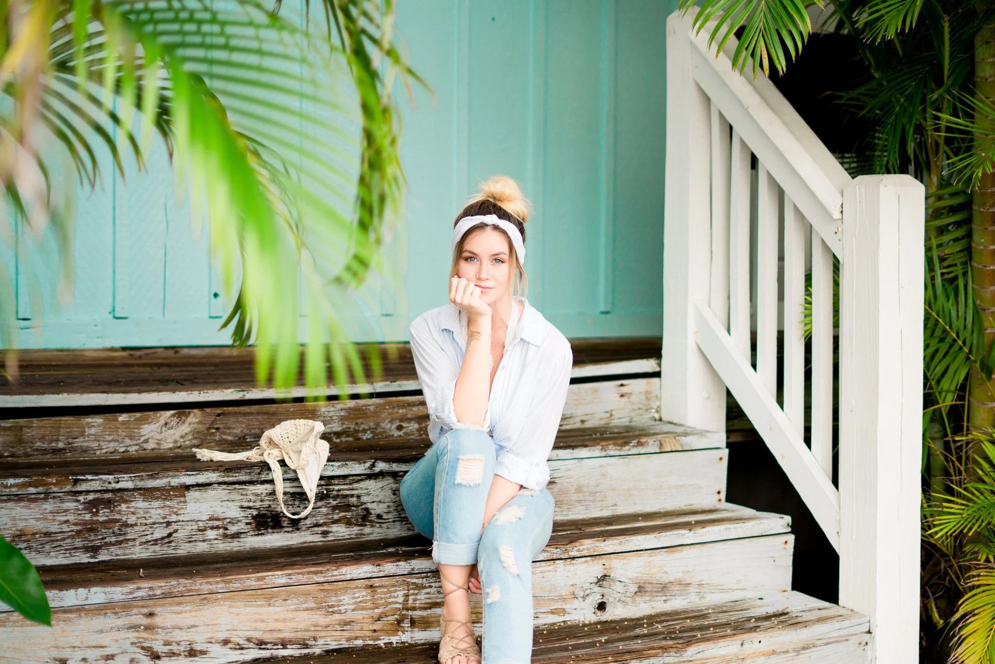 Young woman with blonde hair in a bun, wearing a white headband and white shirt, sitting on weathered wooden steps surrounded by greenery, with a light blue wall in the background. South Florida Portraits.