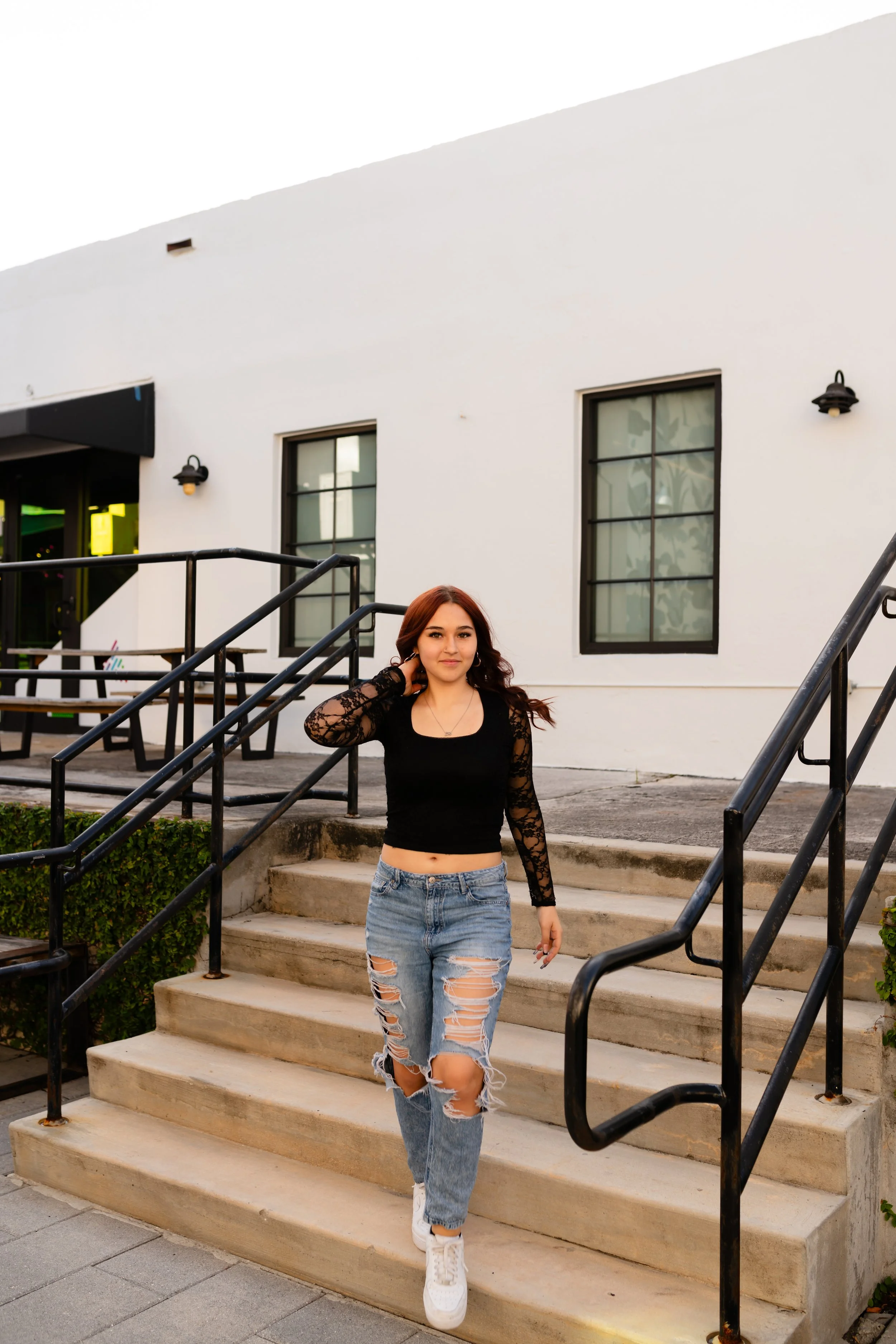 A young woman with red hair, wearing a black lace long-sleeve crop top, ripped jeans, and white sneakers, is walking down concrete stairs outside a white building with black-framed windows.