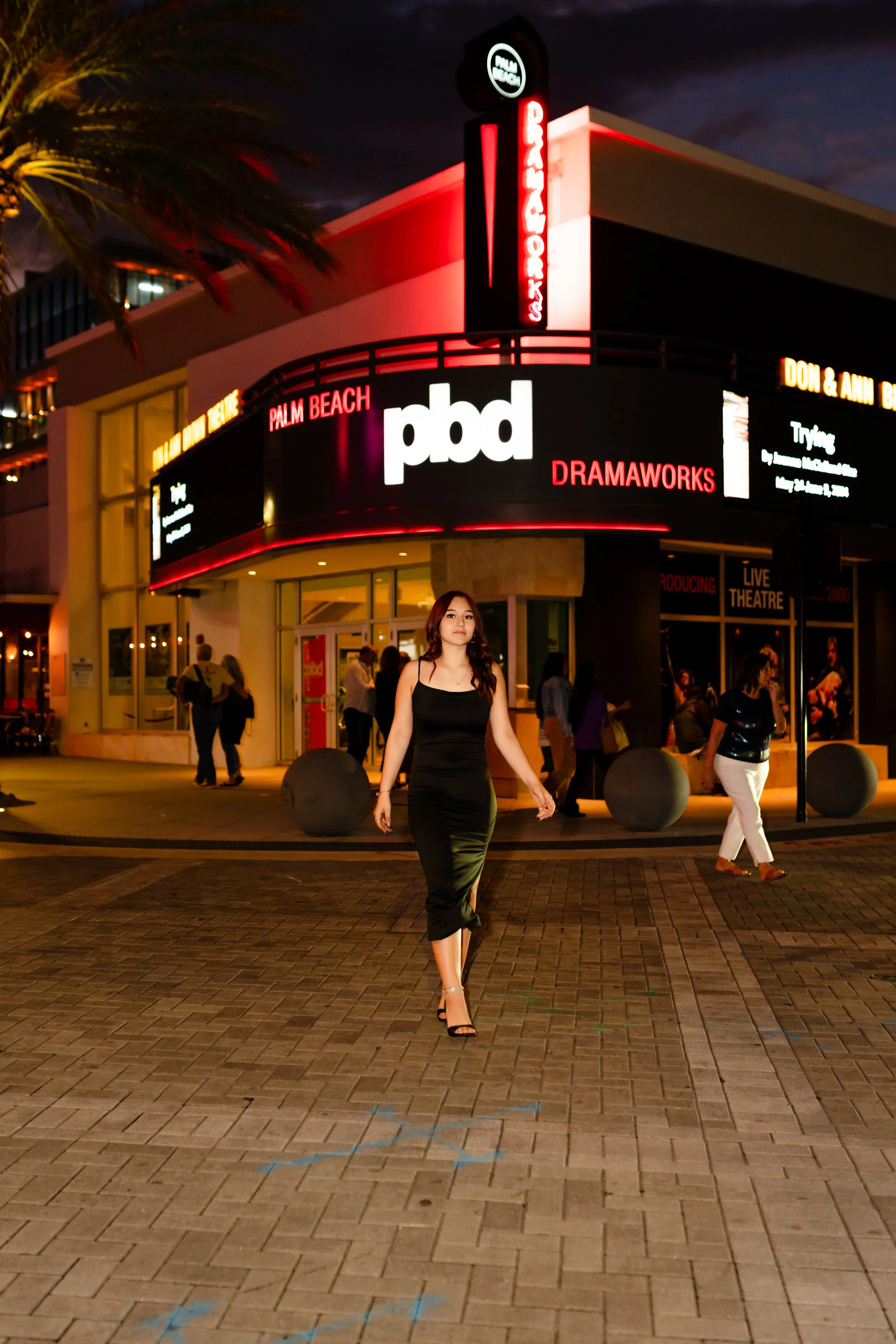 A woman in a black dress walking in front of an illuminated theater marquee at night, with other pedestrians nearby and the marquee displaying "pbd DRAMAWORKS" and other signage for the Palm Beach area.