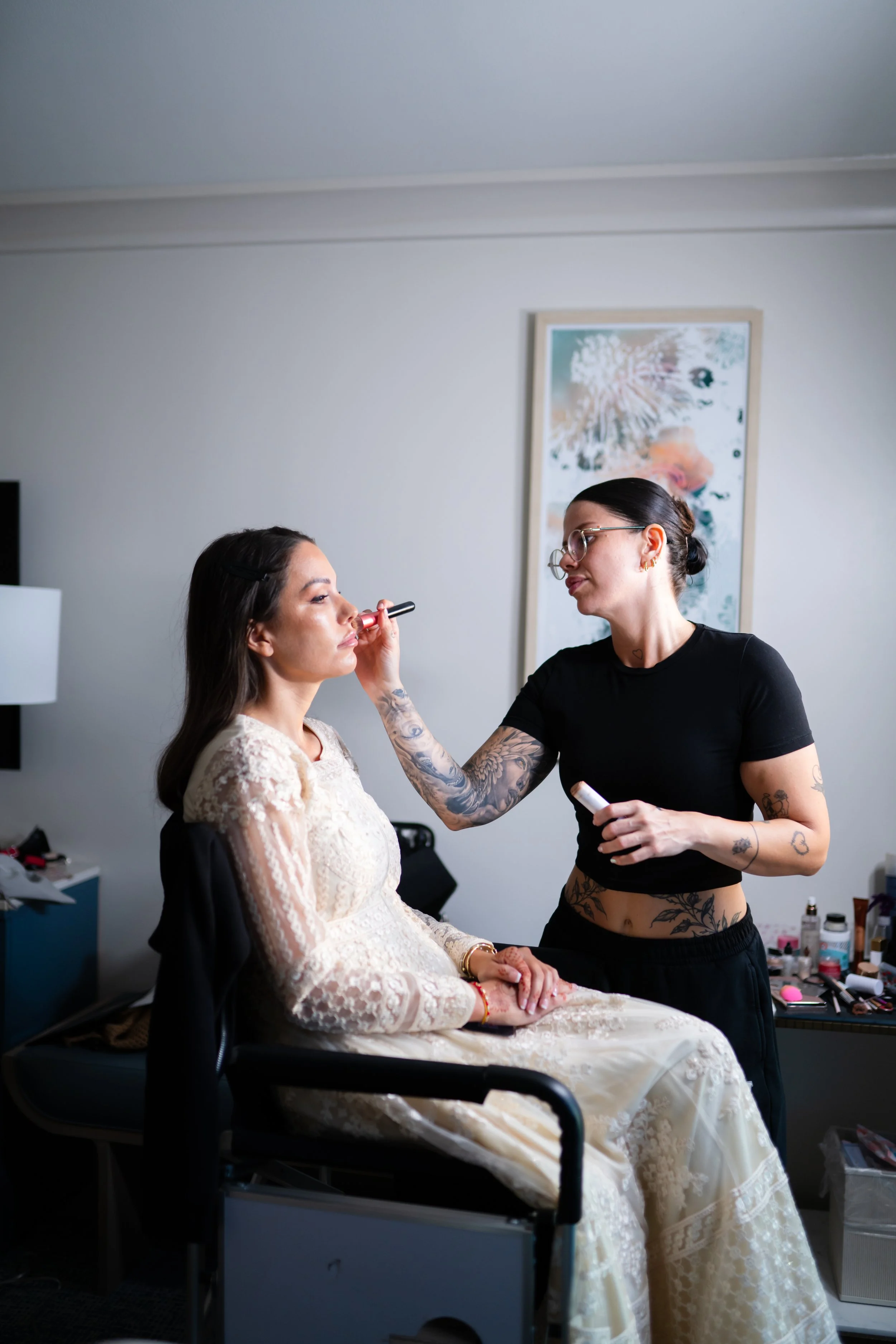 A makeup artist applying makeup to a woman in a white lace dress, seated in a room with various makeup products on a table nearby.