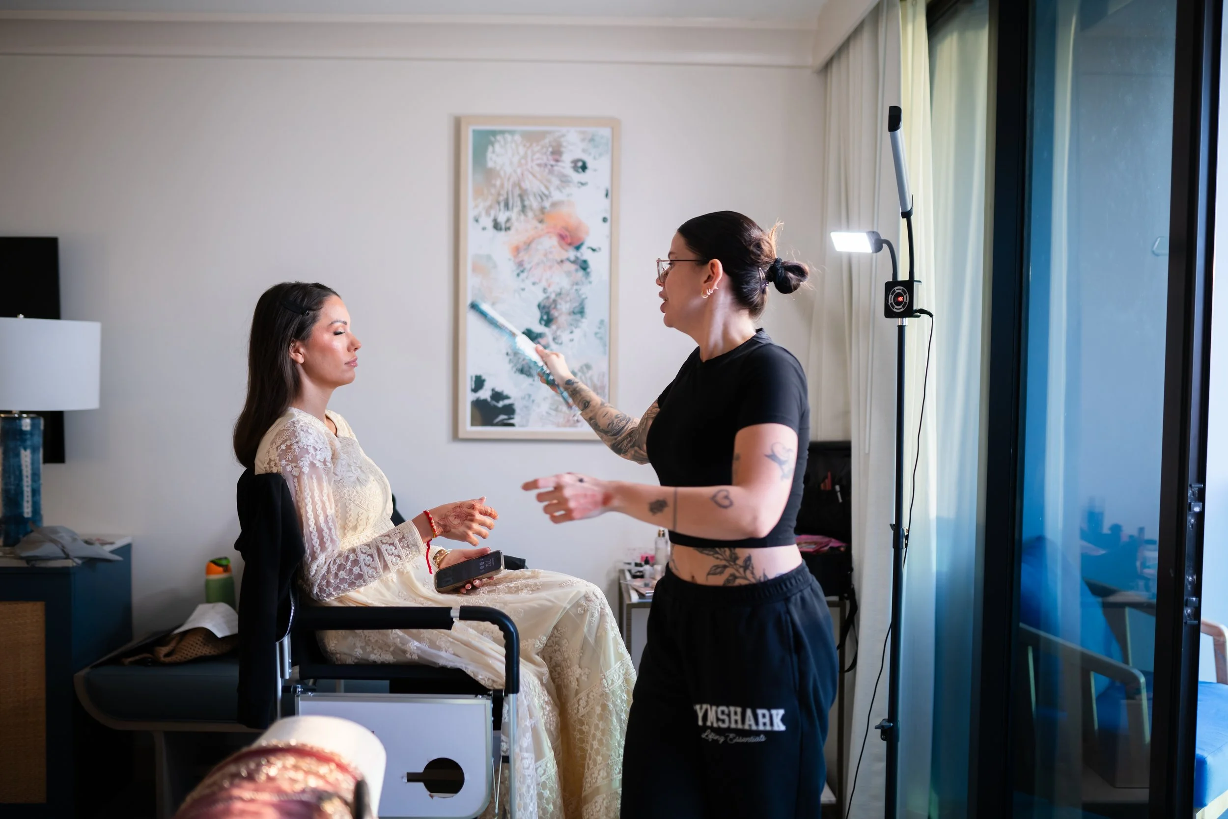 A makeup artist with tattoos applies makeup to a woman in a hospital bed, with natural light coming through sliding glass doors.