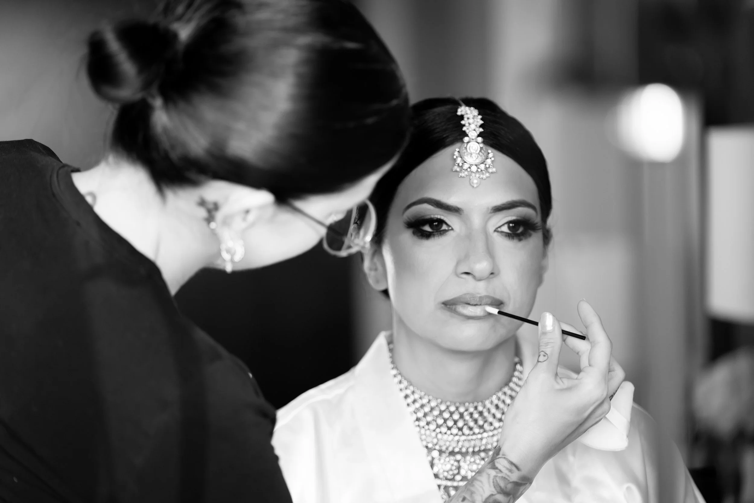 A woman with dark hair and jewelry getting her makeup done by a makeup artist, who is applying lipstick with a brush.