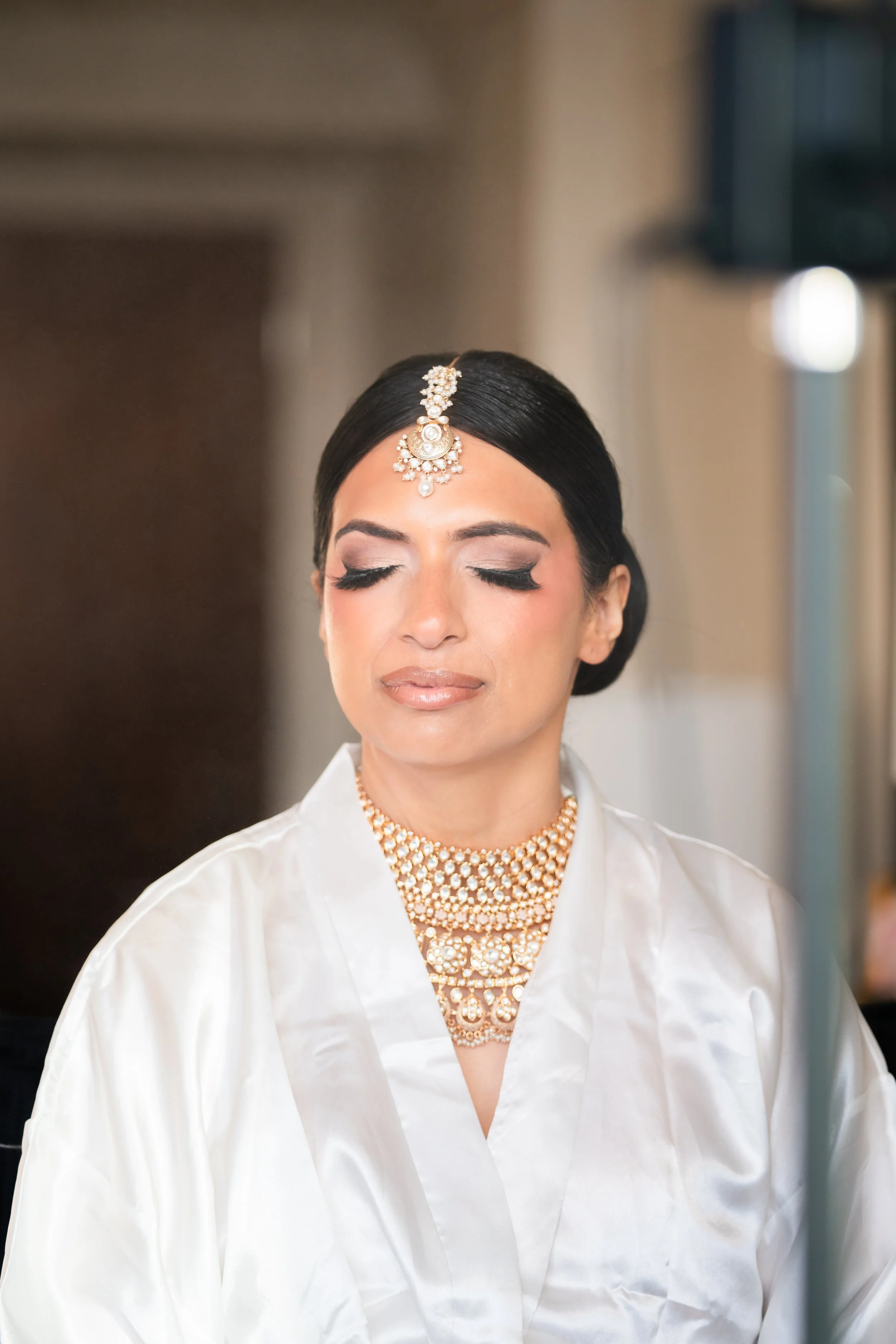 A woman with dark hair styled in an elegant updo, wearing traditional Indian jewelry including a maang tikka on her forehead, a pearl necklace, and matching earrings. She has makeup with smoky eyes and nude lips, dressed in a silk white garment, poss
