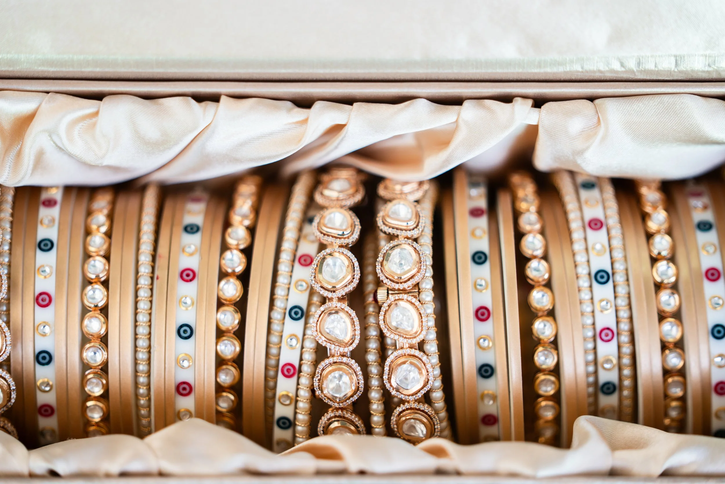 A collection of gold and silver bracelets with various designs, some featuring large clear stones, organized in a jewelry box lined with satin fabric.