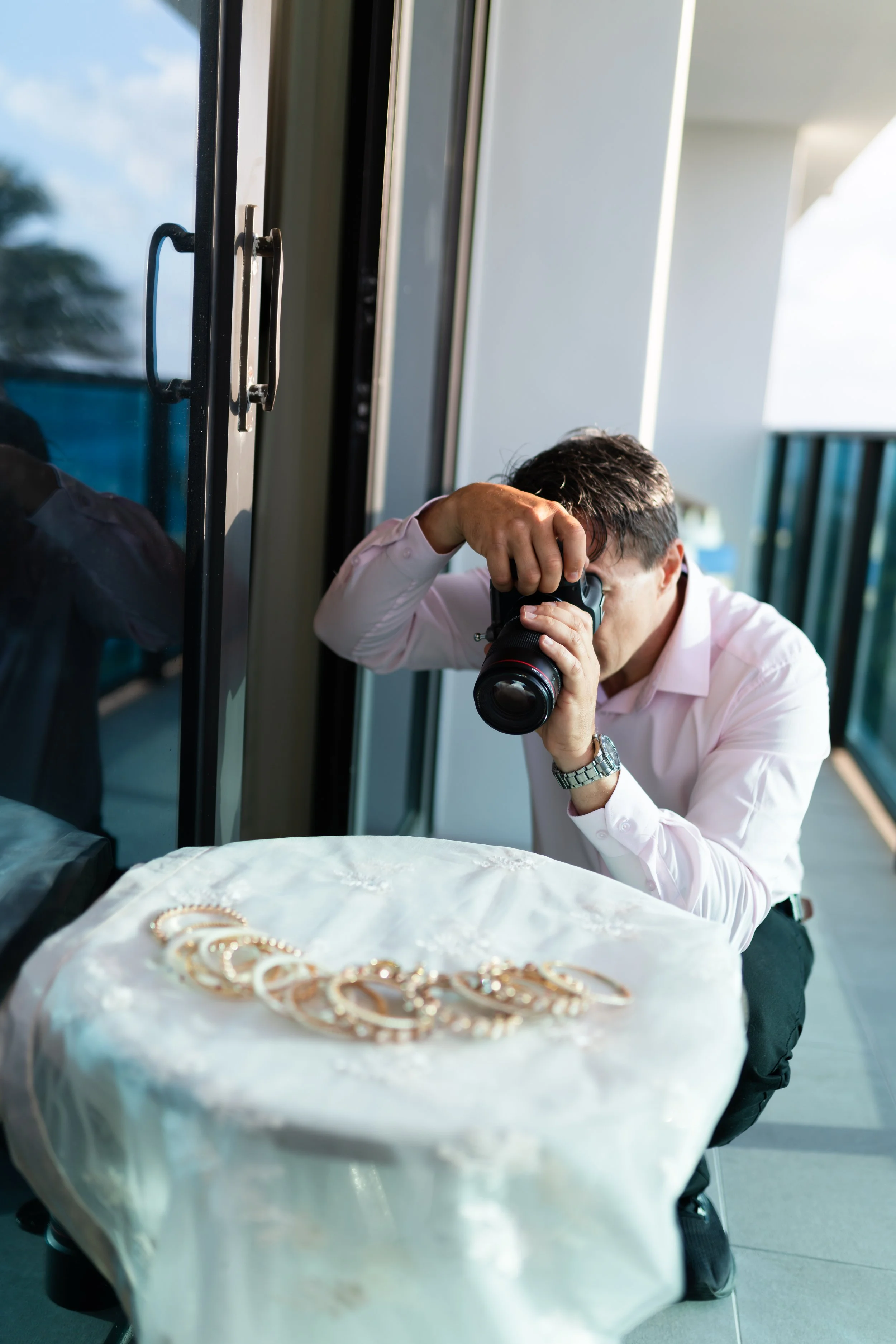 A man in a white shirt takes a photograph of jewelry, possibly earrings, on a table covered with a shiny cloth, on a balcony during daytime.
