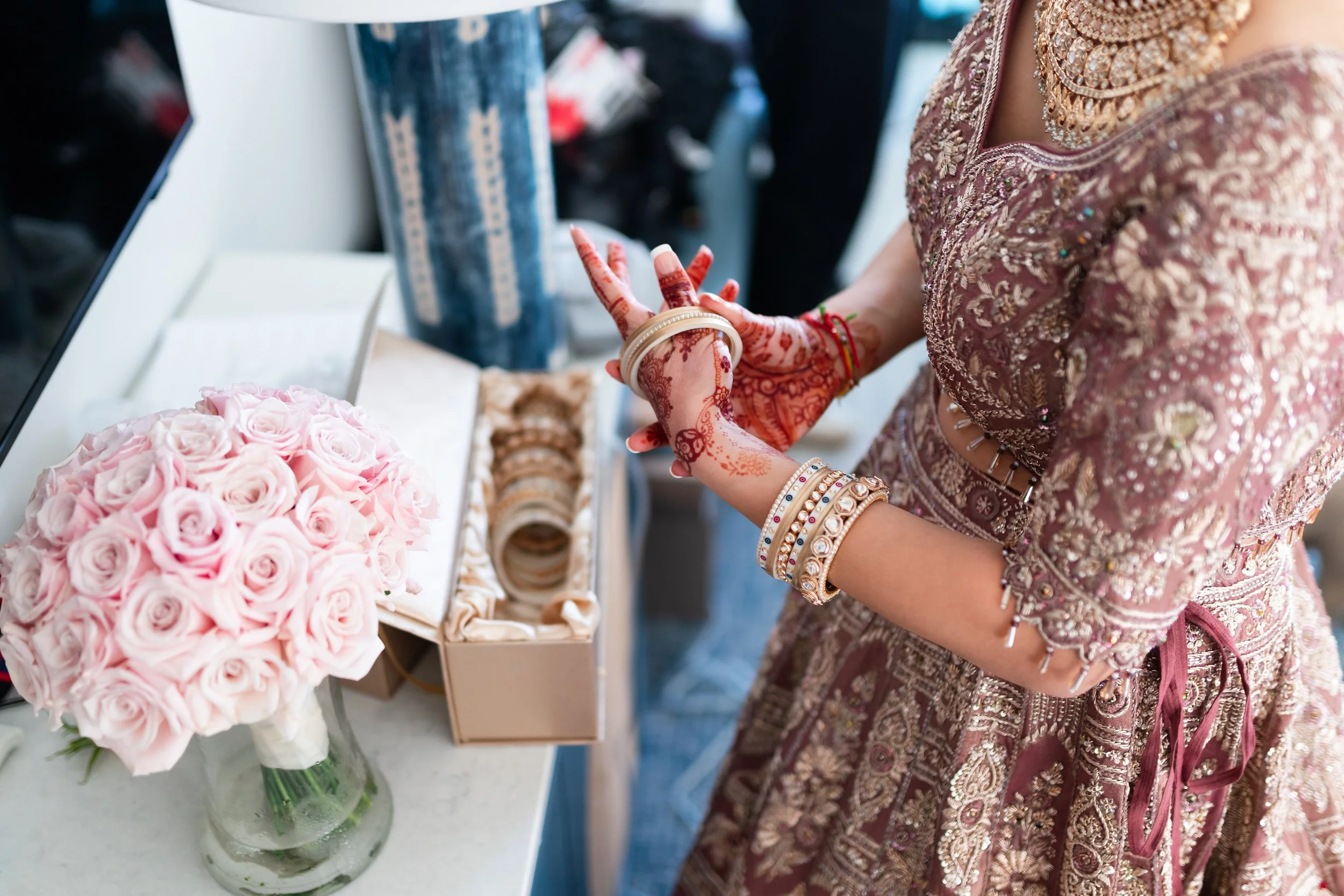 A bride dressed in a richly embroidered mauve and gold traditional Indian outfit is putting on bangles, with watch and jewelry adorning her wrist, near a pink rose bouquet and bangles on display.
