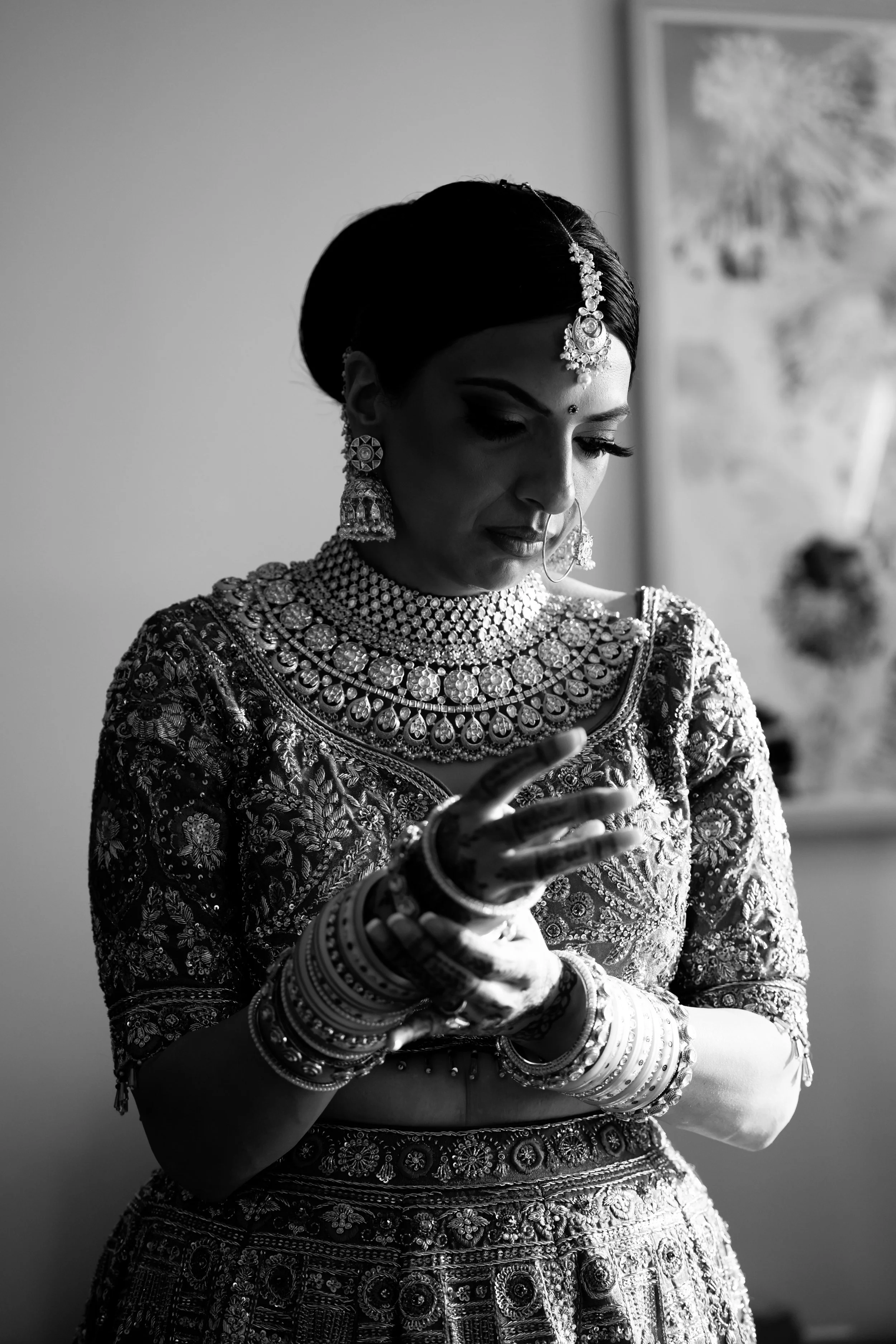 Black and white photo of a woman dressed in traditional Indian bridal attire, wearing elaborate jewelry and bangles, looking at her hand.