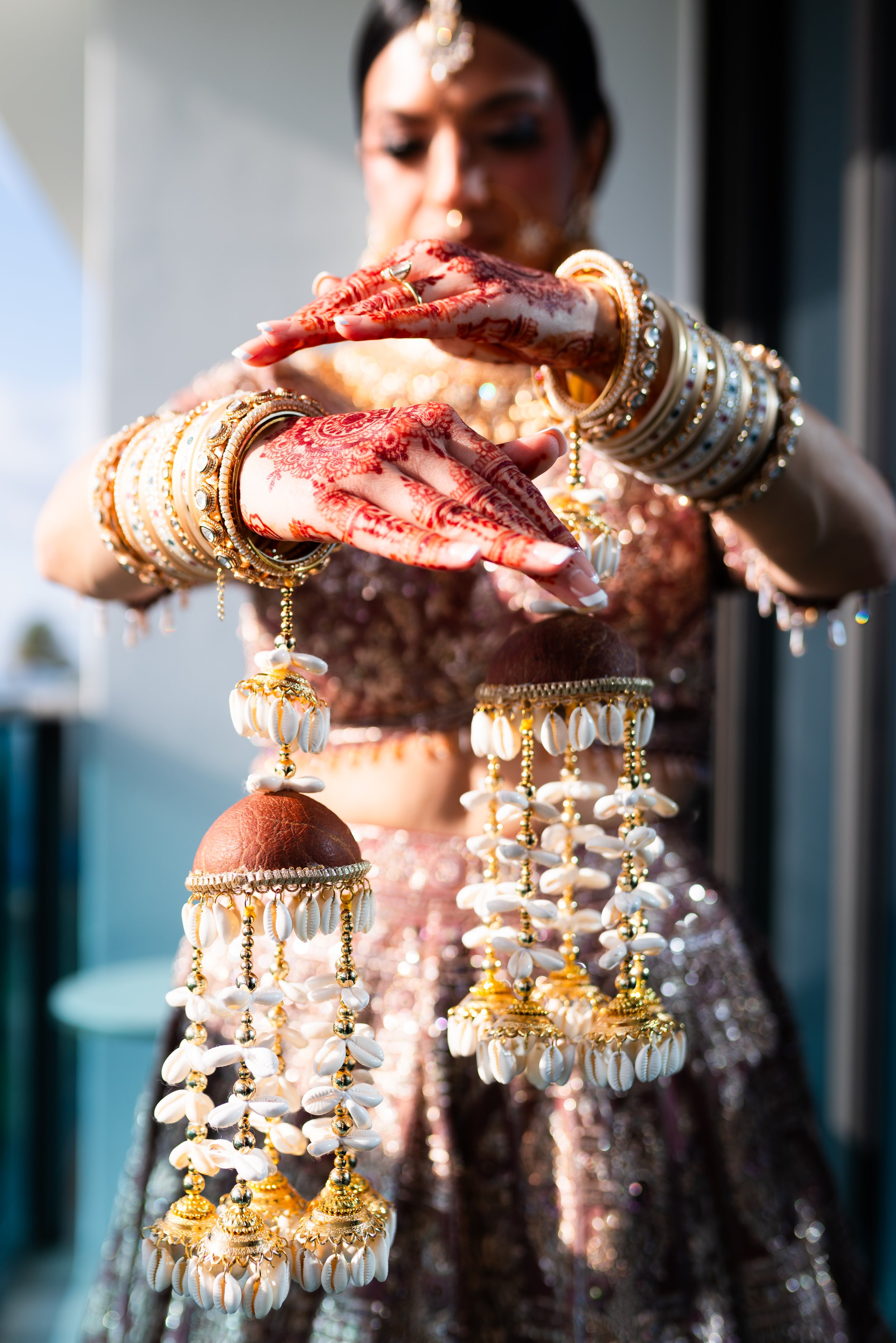 A woman dressed in traditional Indian attire with henna decorated hands, wearing numerous golden bangles and jewelry, holds traditional shell jewelry called 'Jhumkas' in her hands.  South Florida Wedding Photographer, Cali Wedding Photographer